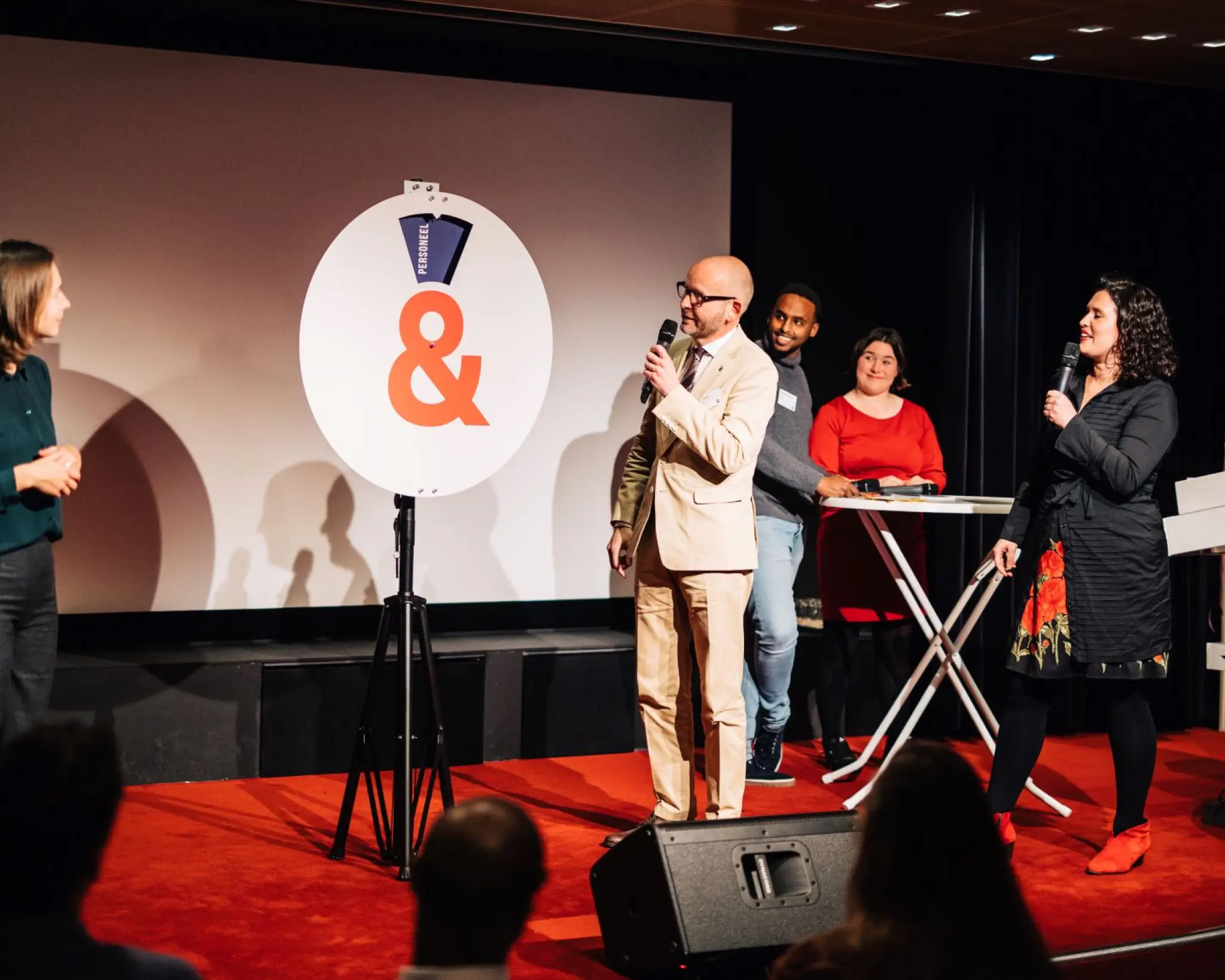 Vijf personen staan op een podium tijdens een bijeenkomst. Twee van hen spreken met een microfoon, terwijl de anderen toekijken. In het midden staat een groot bord met een oranje ampersand.