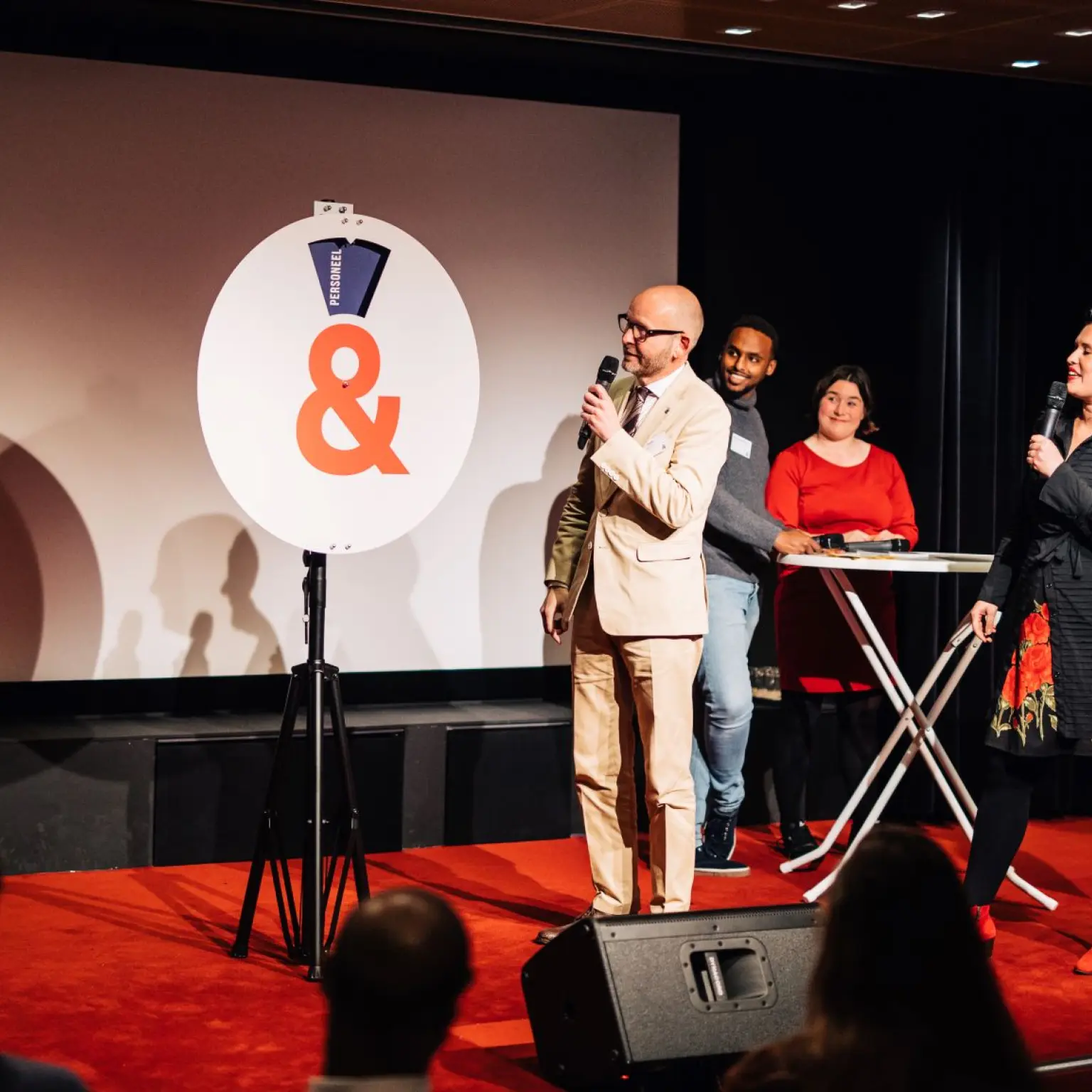 Vijf personen staan op een podium tijdens een bijeenkomst. Twee van hen spreken met een microfoon, terwijl de anderen toekijken. In het midden staat een groot bord met een oranje ampersand.