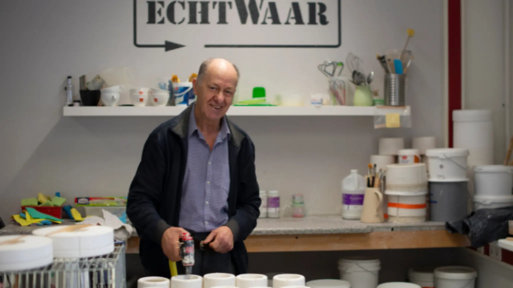 Een man aan de slag in de werkplaats van echtWaar. Om hem heen staan diverse potjes, kwasten, verf en ander materiaal. Op de muur achter hem staat het logo van echtWaar.