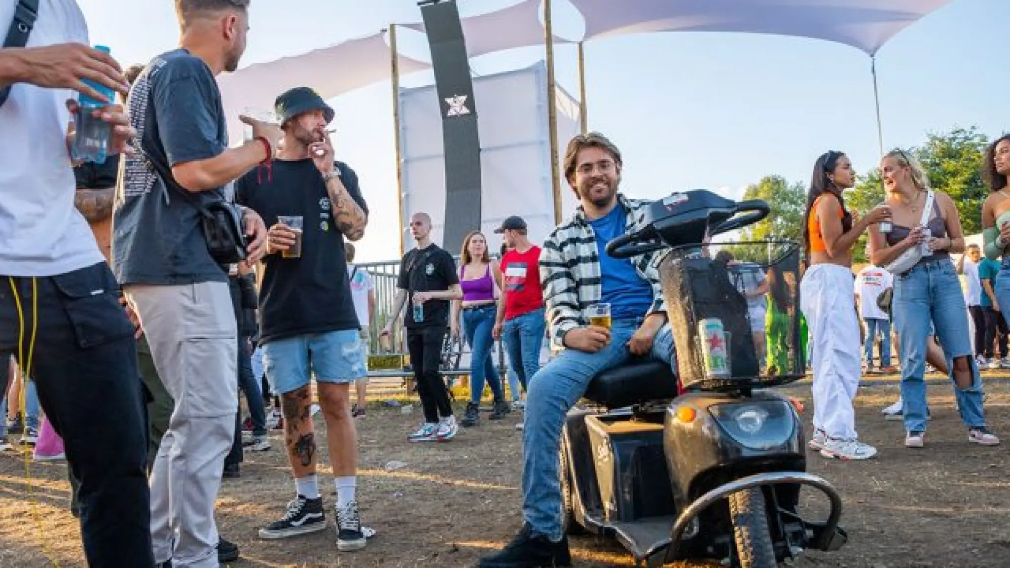 Een jonge man zit op een scootmobiel op een festivalterrein. Hij en andere  festivalgangers om hem heen hebben drankjes in hun hand en genieten van de sfeer.
