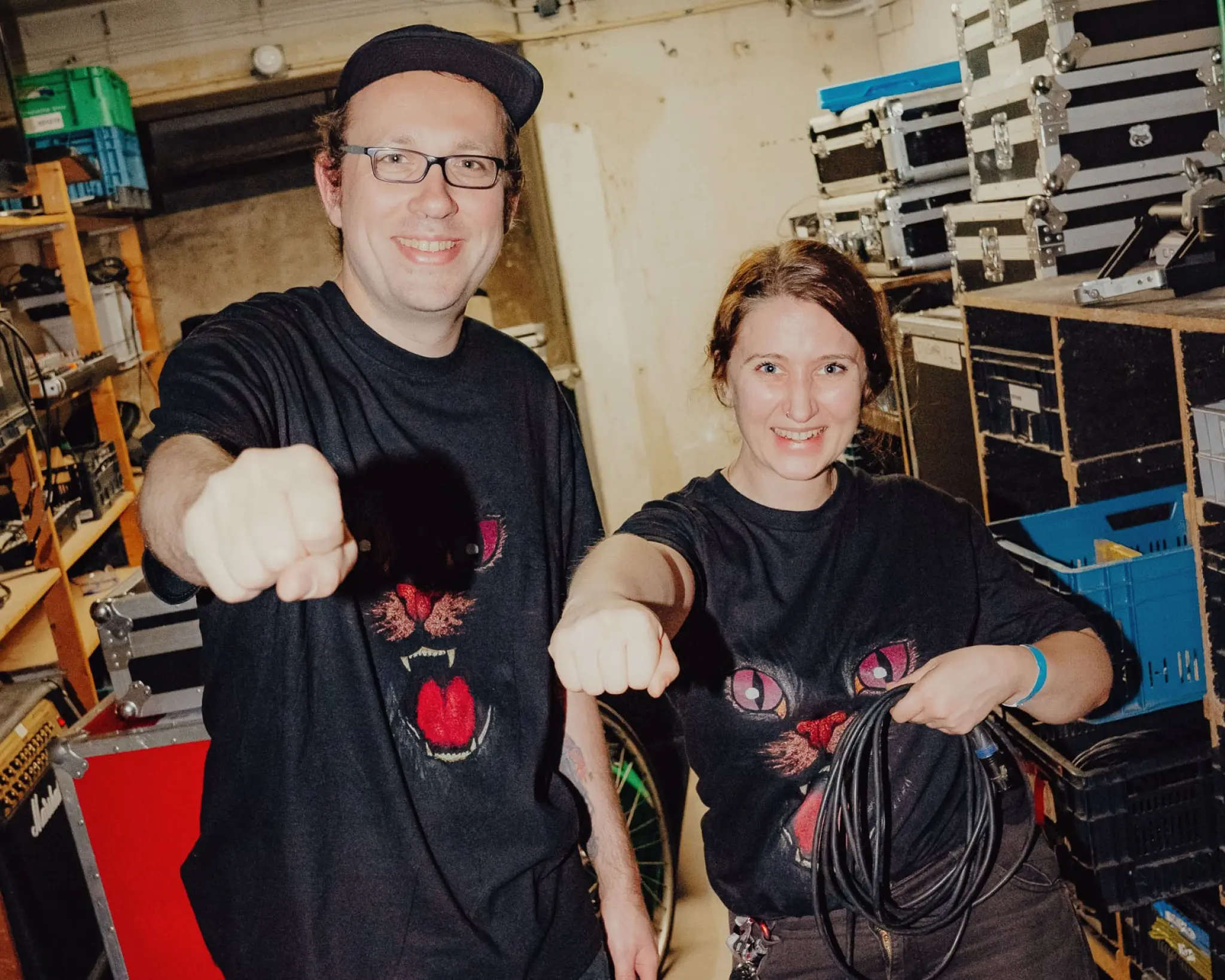Twee mensen in zwarte T-shirts met een kattenprint staan in een opslagruimte vol apparatuur en steken lachend een vuist naar de camera uit.