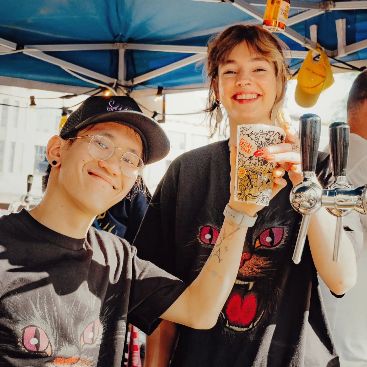 Twee lachende mensen staan achter een dranktap op een evenement en houden samen een beker omhoog. Op de achtergrond staat nog een persoon achter de bar onder een blauwe partytent.