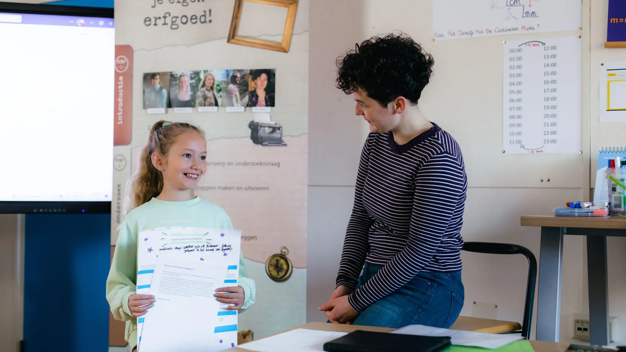 Foto van Anke Spieringhs naast een kind in een klaslokaal. Het kind lacht en houdt presentatiemateriaal vast. Foto door Fabia Cnossen.