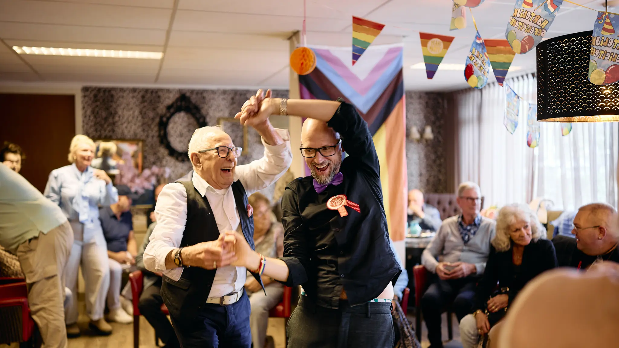 Twee mensen dansen samen en houden elkaars handen vast in een ruimte met andere aanwezigen. Op de achtergrond hangen regenboogvlaggen en feestelijke versiering.