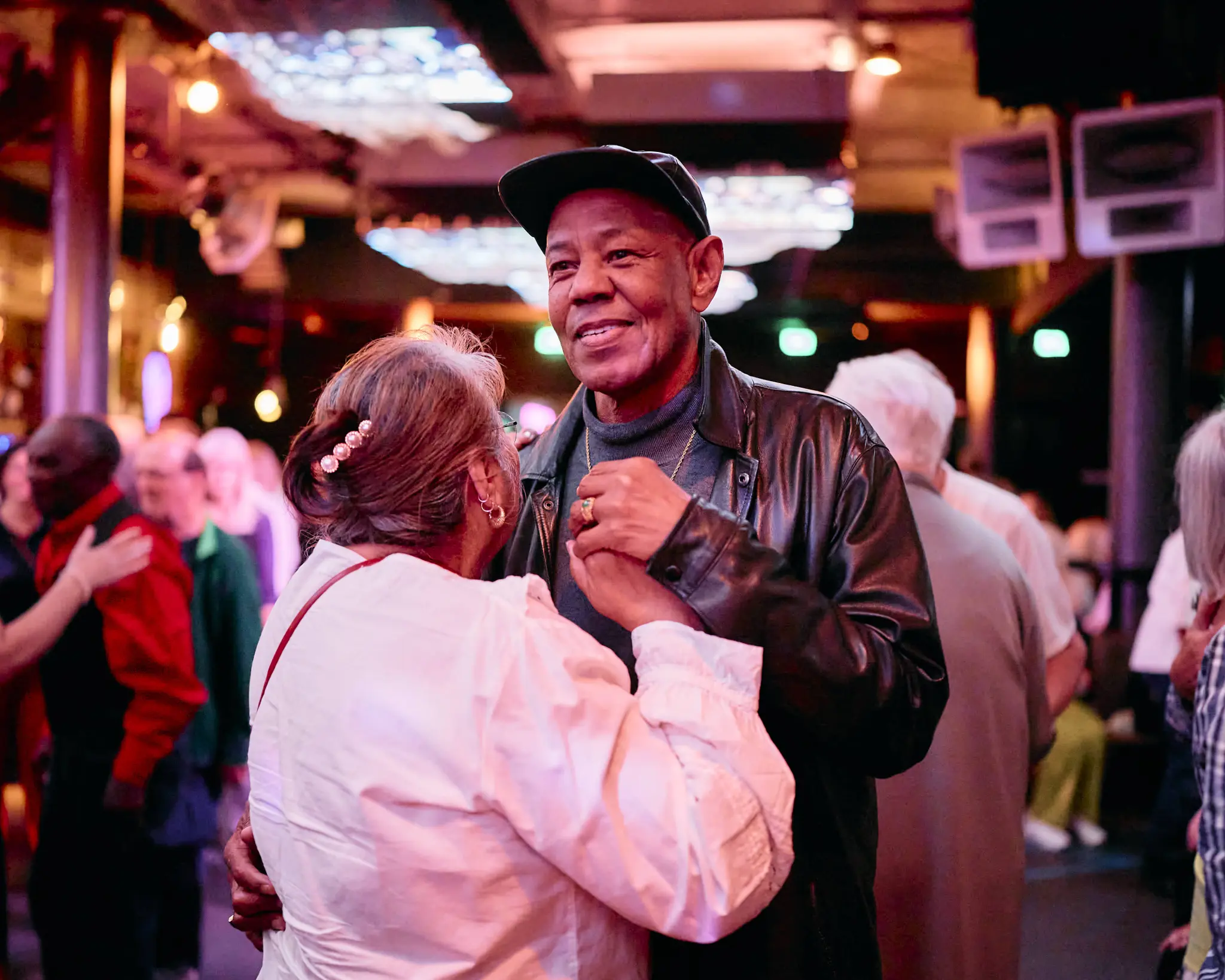 Twee mensen dansen samen in een ruimte met warme, gekleurde verlichting. Op de achtergrond zijn meerdere andere mensen aan het dansen en met elkaar in gesprek.