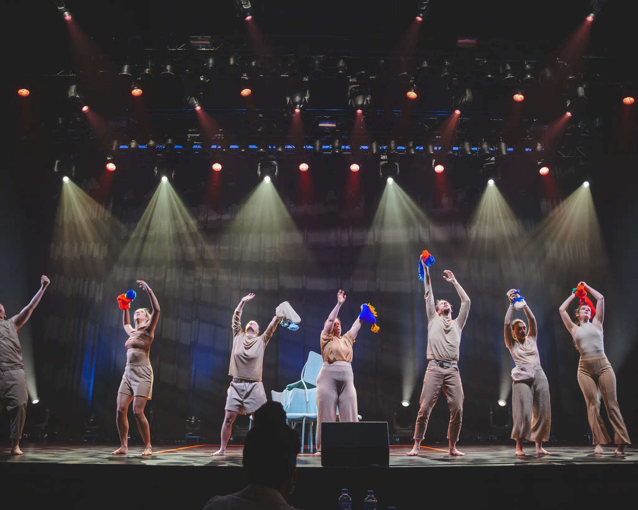 Zeven performers staan op een verlicht podium en houden kleurrijke handpoppen en doeken omhoog tijdens een gezamenlijke voorstelling.