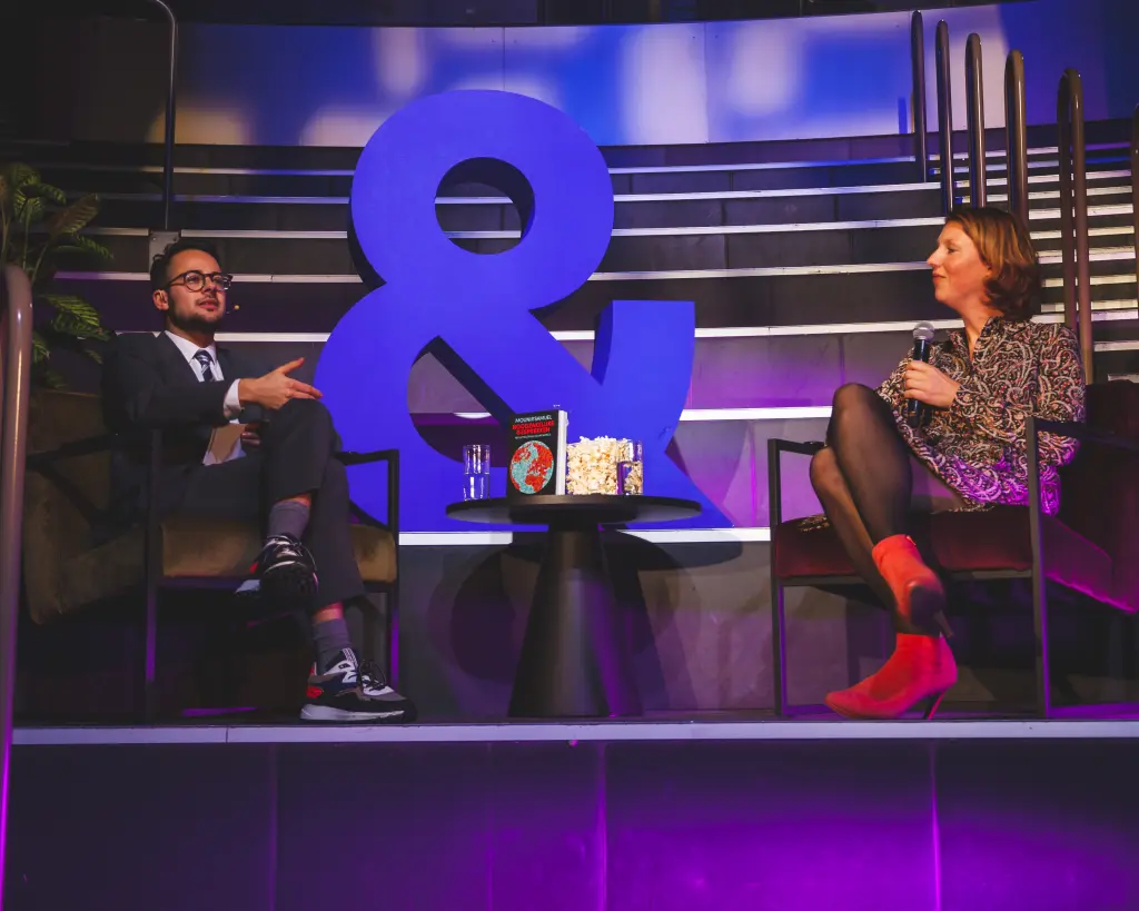 Twee personen zitten tegenover elkaar op een podium en voeren een gesprek met microfoons. Tussen hen staat een groot blauw ampersand, met op de achtergrond een verlichte tribune.