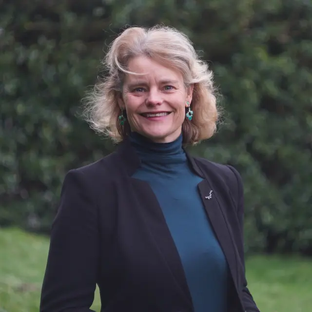 Portret van een glimlachende Mare de Groot, met halflang blond haar, gekleed in een donker jasje en een blauw coltruitje, staand buiten met bomen op de achtergrond.