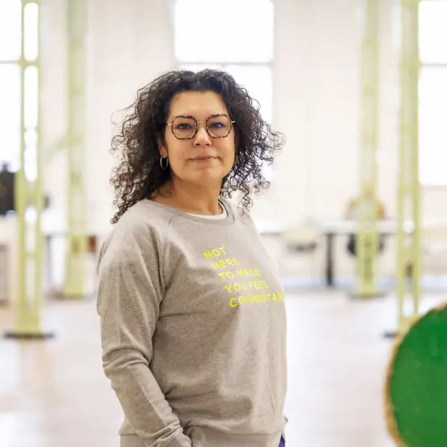 Seyda staat in een grote industriële en lichte ruimte en kijkt neutraal naar de camera. Ze draagt een grijze trui met de tekst 'not here to make you feel comfortable'.
