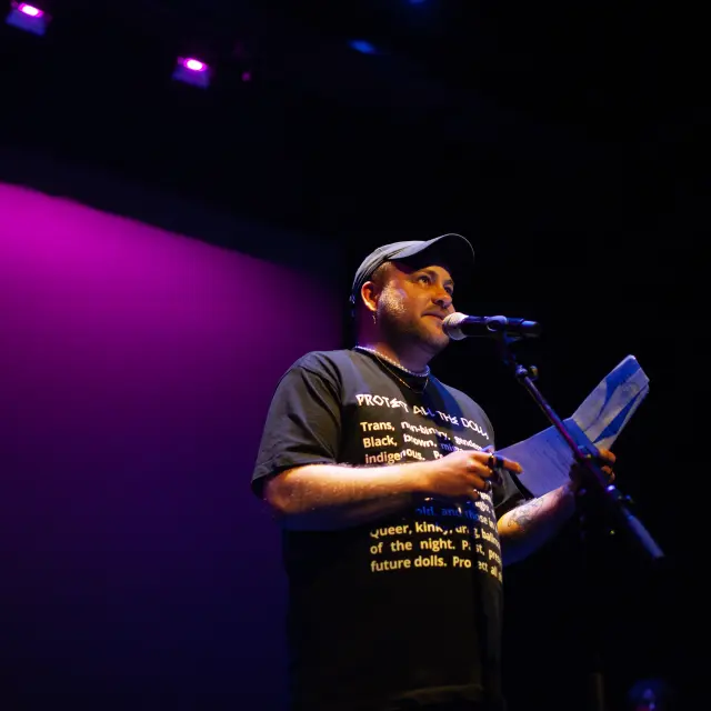 Tijn de Jong staat op een podium, spreekt in een microfoon en houdt enkele papieren vast. Het podium wordt verlicht door paars en blauw theaterlicht, met een donkere achtergrond.