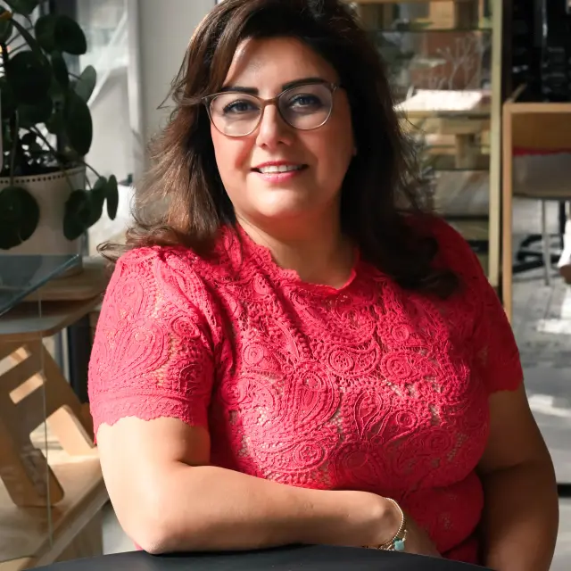 Samira Barari, gekleed in een rood shirt, zit in een lichte kantoorruimte aan een zwarte ronde tafel en kijkt in de camera.