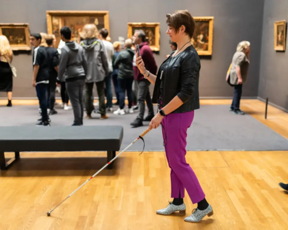 Een persoon met een witte taststok gebruikt een smartphone in een museumzaal met schilderijen aan de muur, terwijl andere bezoekers de kunst bekijken.