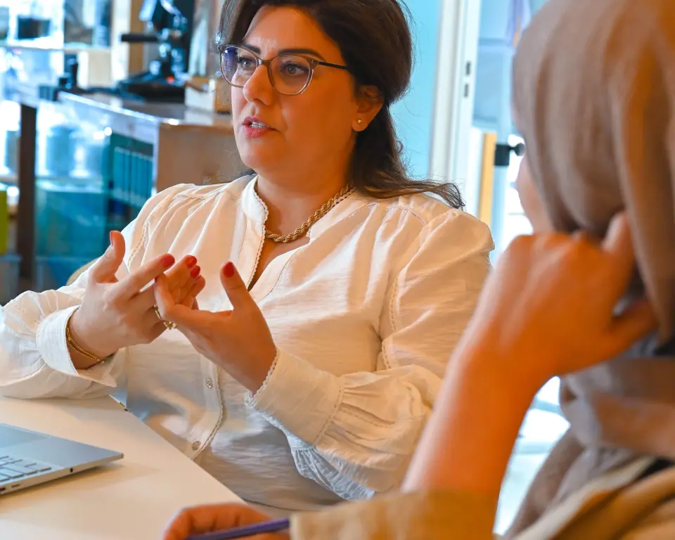 Samira Barari, gekleed in een witte blouse, legt iets uit tijdens een gesprek aan tafel, terwijl een andere persoon aantekeningen maakt in een lichte werkruimte.