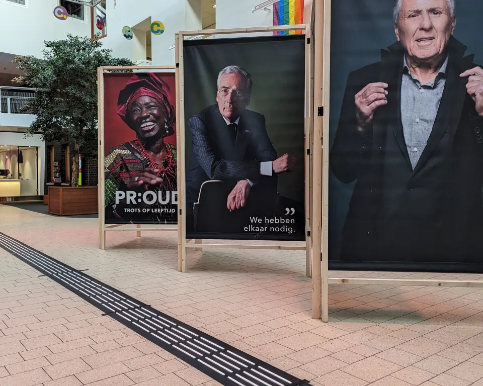 Tijdelijke expositie van grote portretfoto's op houten borden. Ervoor langs loopt een geleidelijn op die met behulp van matten op de vloer is aangevracht.