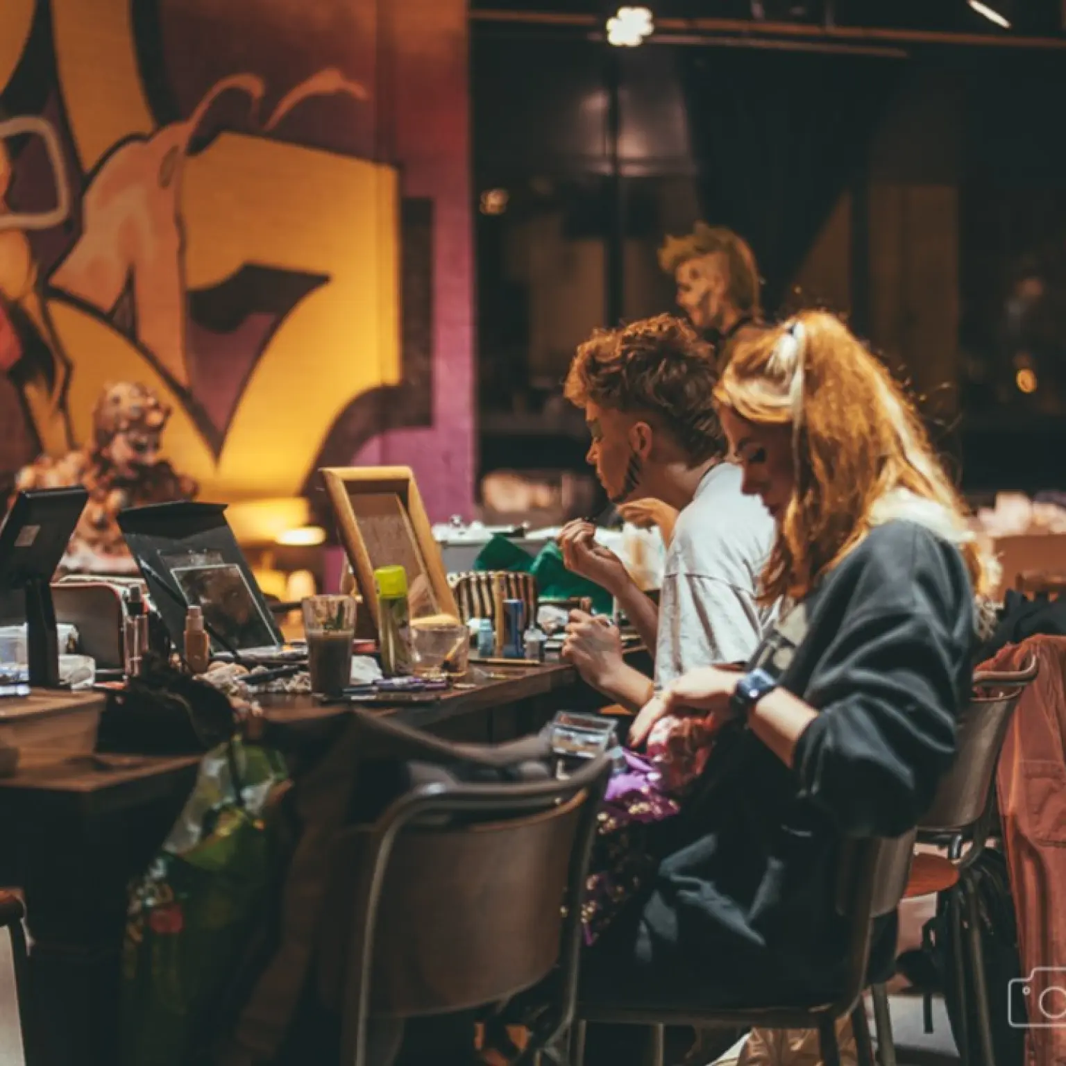 Meerdere personen zitten aan tafels in een sfeervol verlichte ruimte en bereiden zich voor achter spiegels en make-upmaterialen. Op de achtergrond is een grote muurschildering zichtbaar.