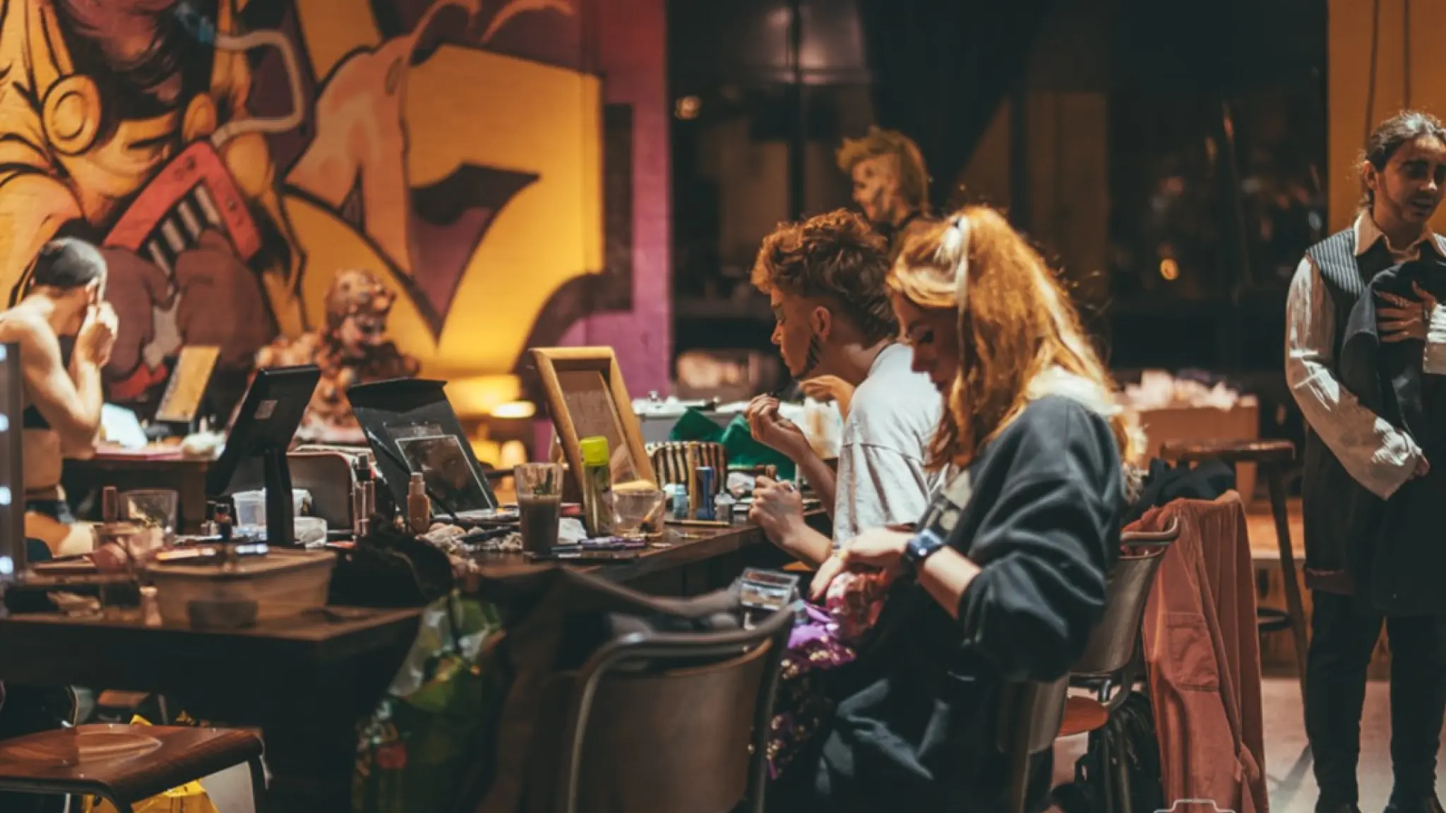 Meerdere personen zitten aan tafels in een sfeervol verlichte ruimte en bereiden zich voor achter spiegels en make-upmaterialen. Op de achtergrond is een grote muurschildering zichtbaar.