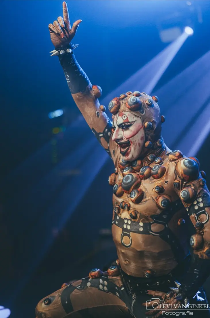 Persoon in een opvallend podiumkostuum met tientallen oogvormige elementen op het lichaam, staand onder blauwe spotlights met één arm omhoog gestoken tijdens een podiumoptreden.