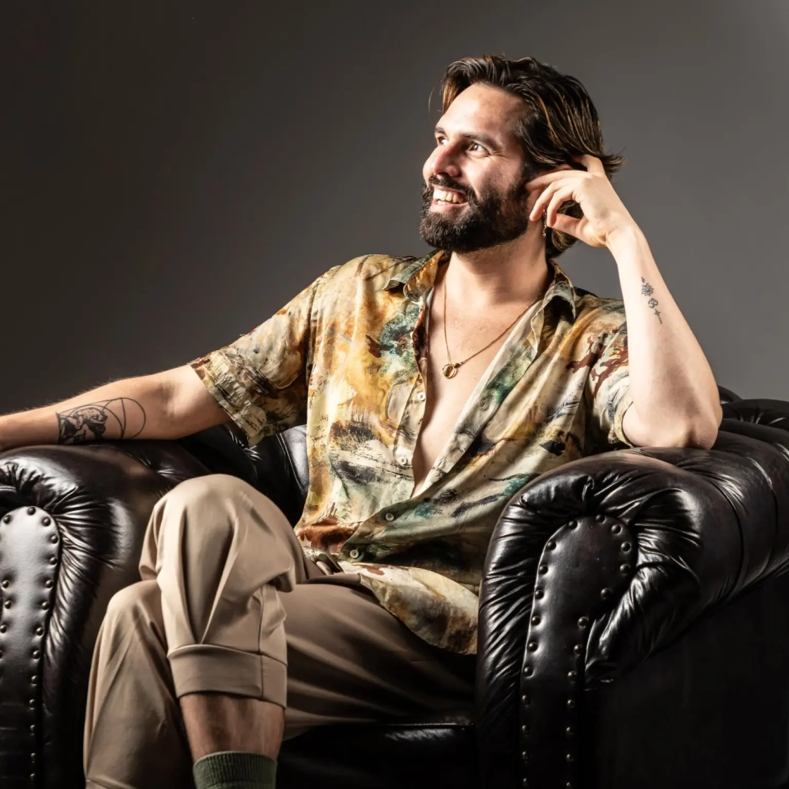 Luis Bracamontes zit in een donkere leren fauteuil. Hij leunt met zijn hoofd op zijn linkerhand en kijkt lachend naar rechts, schuin weg van de camera. Zijn blouse hangt half open en hij draagt een ketting. Op zijn onderarmen zijn tatoeages te zien.