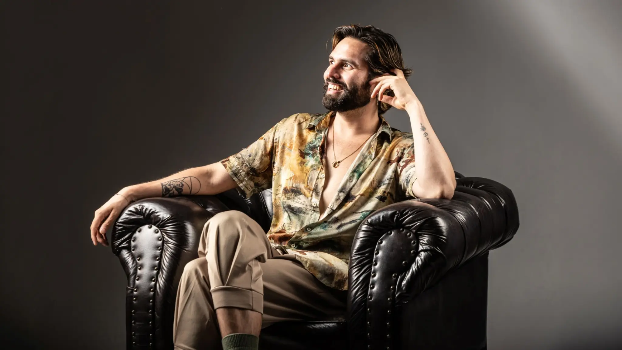 Luis Bracamontes zit in een donkere leren fauteuil. Hij leunt met zijn hoofd op zijn linkerhand en kijkt lachend naar rechts, schuin weg van de camera. Zijn blouse hangt half open en hij draagt een ketting. Op zijn onderarmen zijn tatoeages te zien.