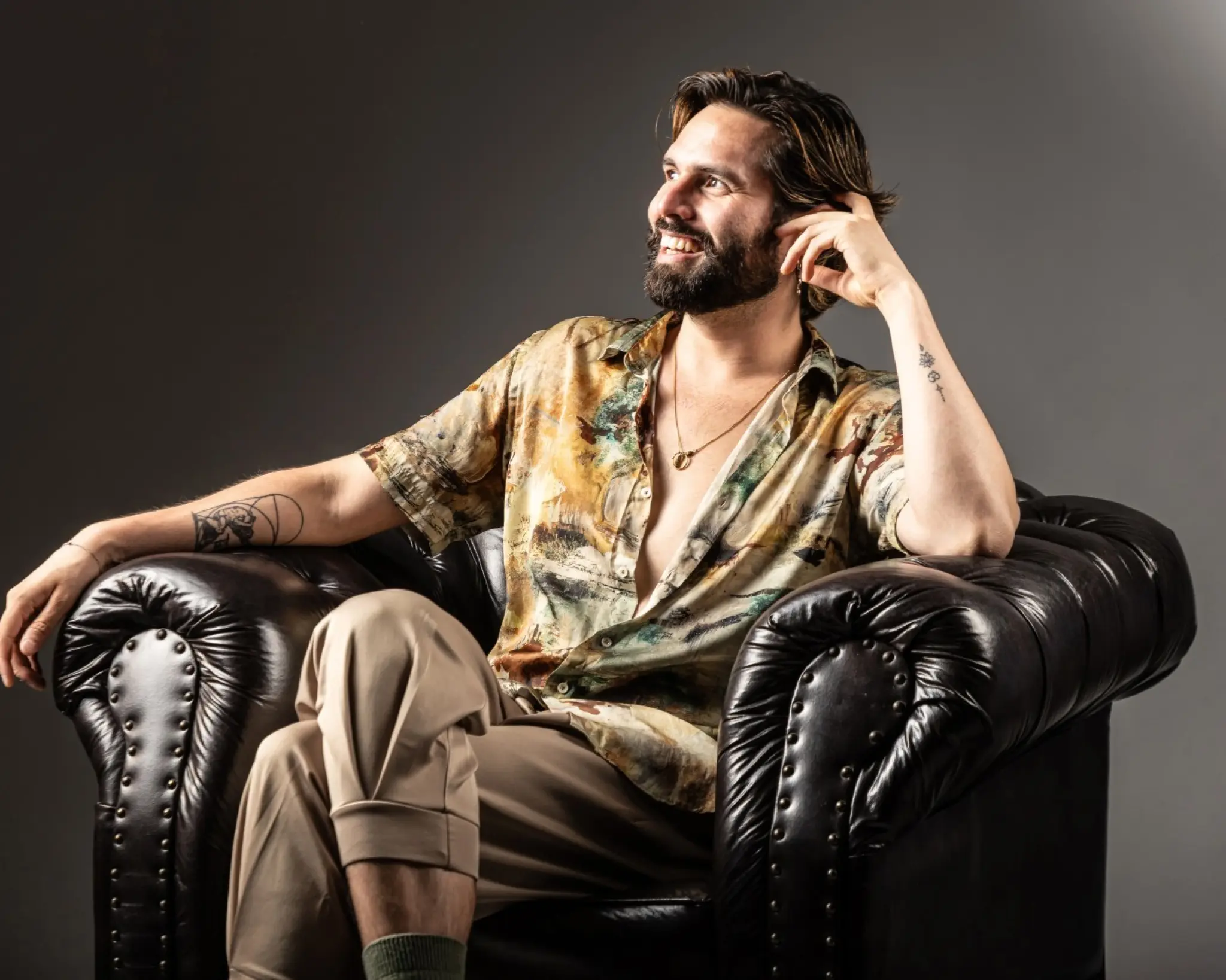 Luis Bracamontes zit in een donkere leren fauteuil. Hij leunt met zijn hoofd op zijn linkerhand en kijkt lachend naar rechts, schuin weg van de camera. Zijn blouse hangt half open en hij draagt een ketting. Op zijn onderarmen zijn tatoeages te zien.