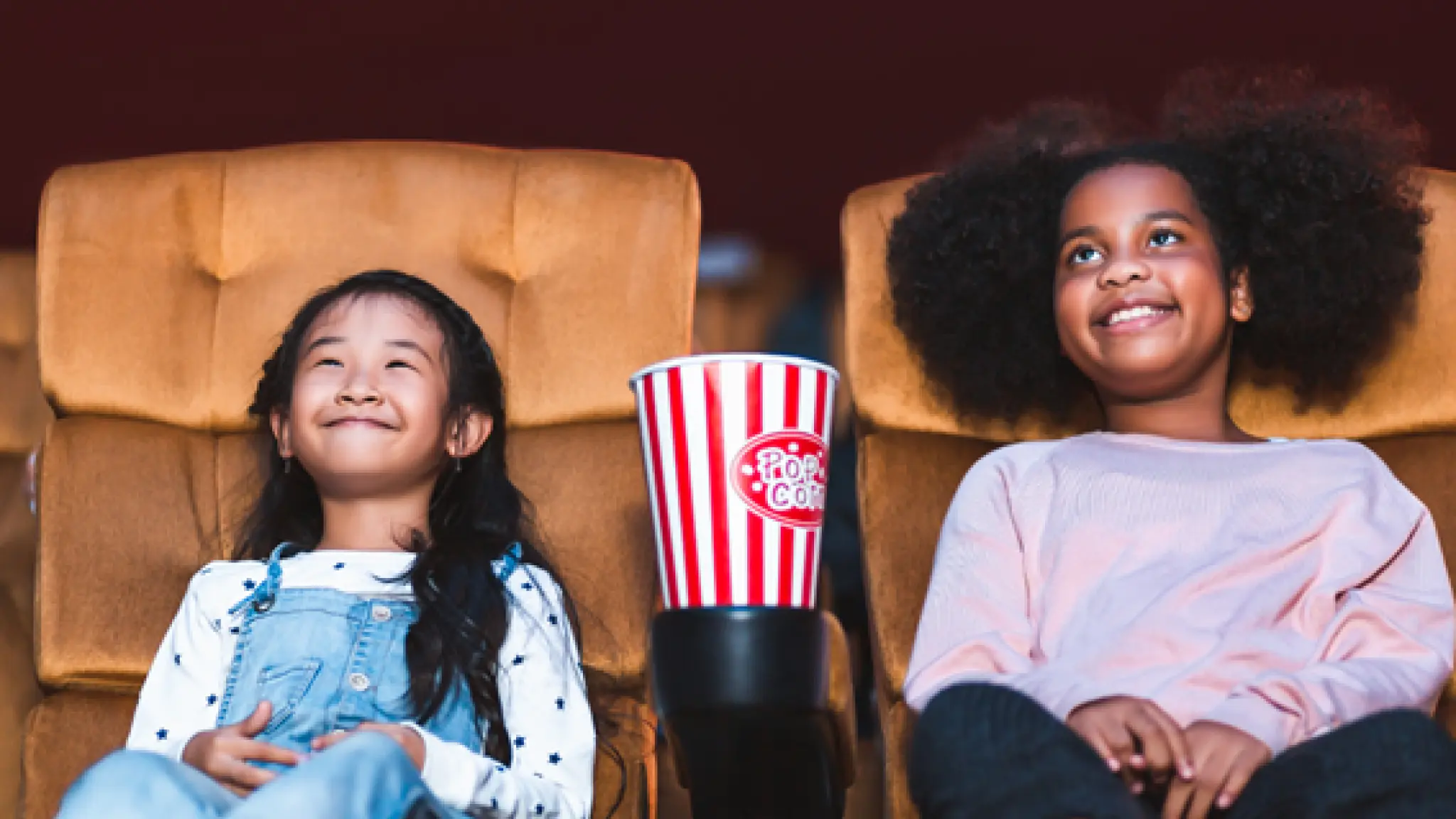 Een meisje met Aziatisch uiterlijk en een meisje met Afrikaans uiterlijk zitten in bioscoopstoelen met een beker popcorn tussen hen in.