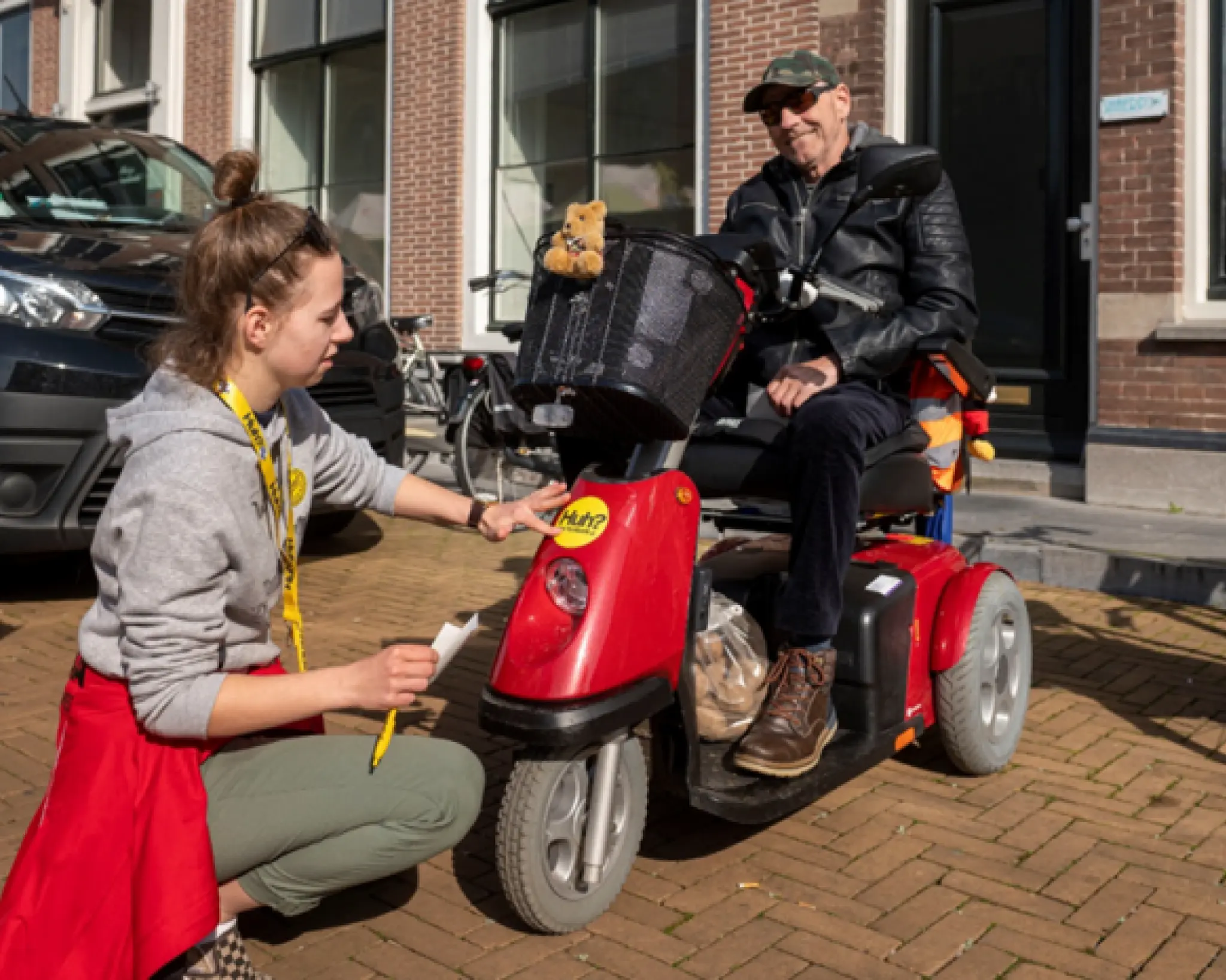 Een jonge vrouw plakt een Huh?-sticker op de scootmobiel van een oudere man.