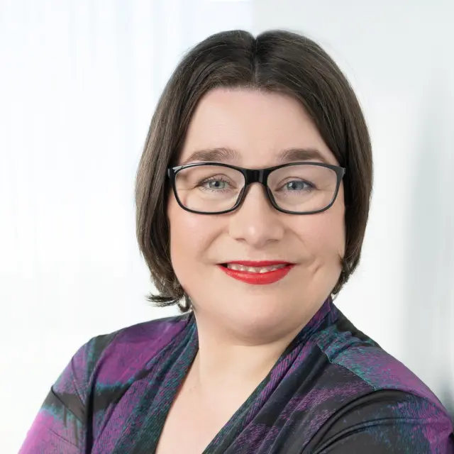 Marianne Dijkshoorn. Ze leunt tegen een witte muur en kijkt over haar linkerschouder naar de camera.