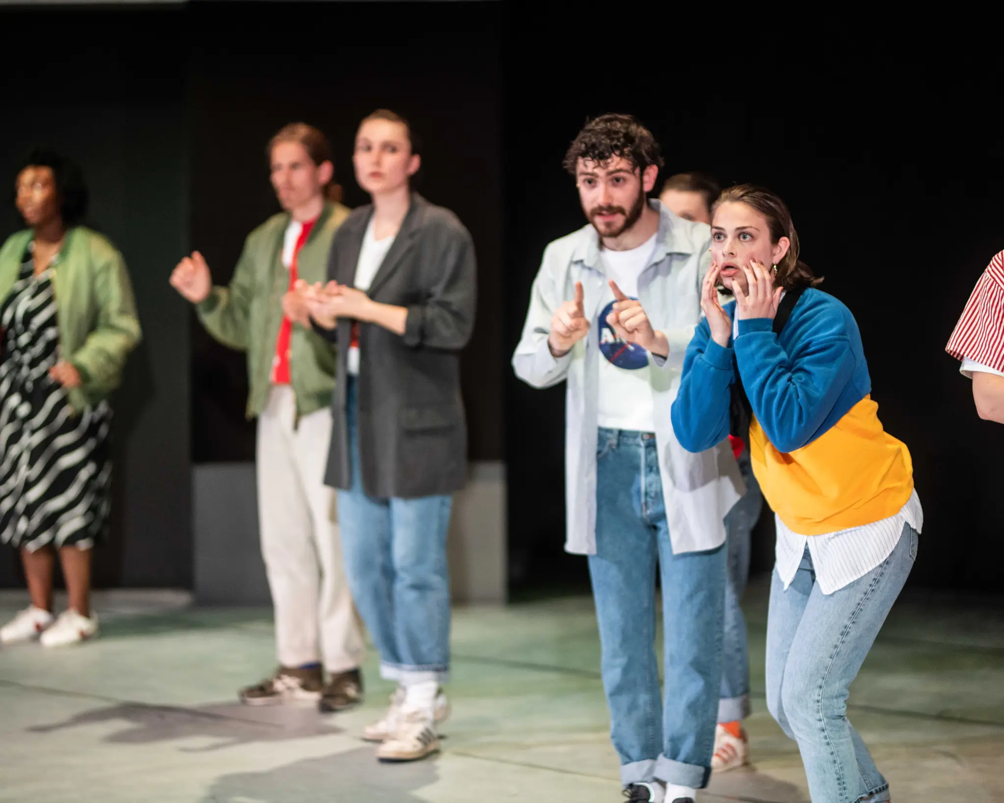 Een groep performers staat op een theaterpodium tijdens een scène. Op de voorgrond reageert een persoon in een blauw-gele trui zichtbaar geschrokken, met de handen tegen het gezicht. Daarnaast gebaart een andere speler met opgestoken wijsvingers. Op de achtergrond staan meerdere performers verspreid over het podium, terwijl de aandacht gericht lijkt op iets buiten beeld.