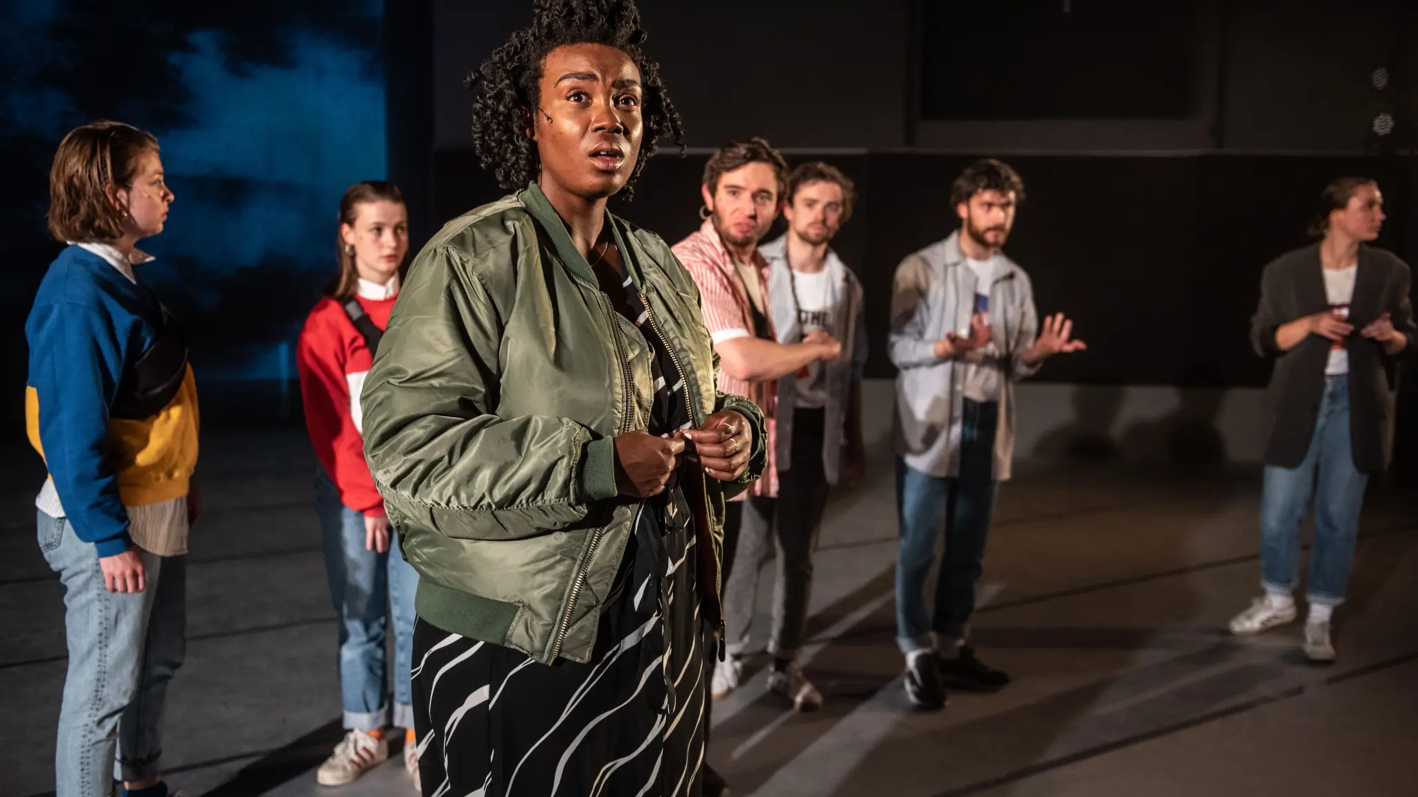 Een groep van zeven performers staat op een theaterpodium. Op de voorgrond kijkt een persoon in een groene bomberjas en een zwart-witte jurk met een verbaasde uitdrukking naar voren. Op de achtergrond staan andere spelers verspreid over het podium, deels onscherp. Het toneel is schaars ingericht en wordt verlicht met dramatisch theaterlicht.