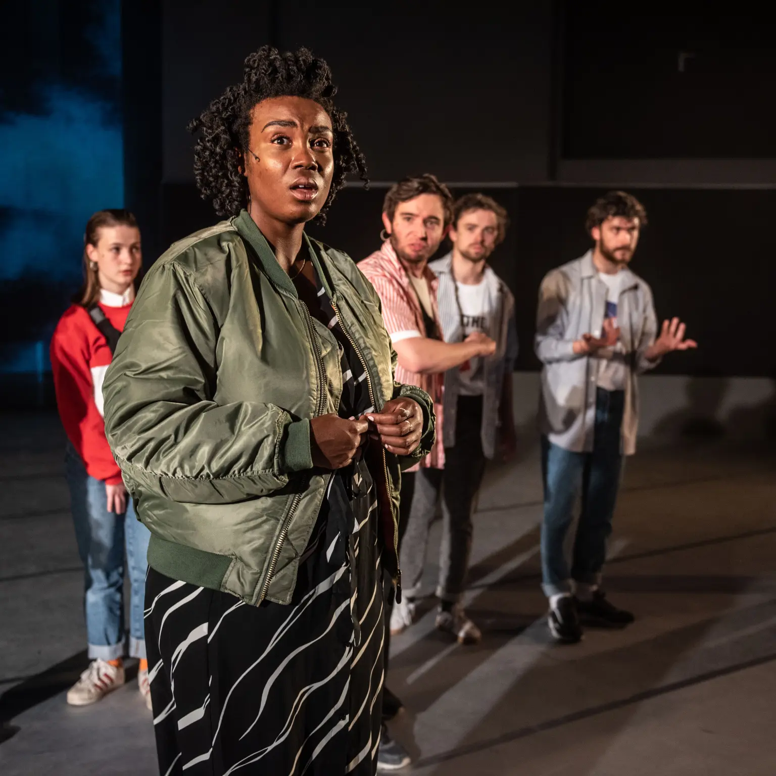 Een groep van zeven performers staat op een theaterpodium. Op de voorgrond kijkt een persoon in een groene bomberjas en een zwart-witte jurk met een verbaasde uitdrukking naar voren. Op de achtergrond staan andere spelers verspreid over het podium, deels onscherp. Het toneel is schaars ingericht en wordt verlicht met dramatisch theaterlicht.