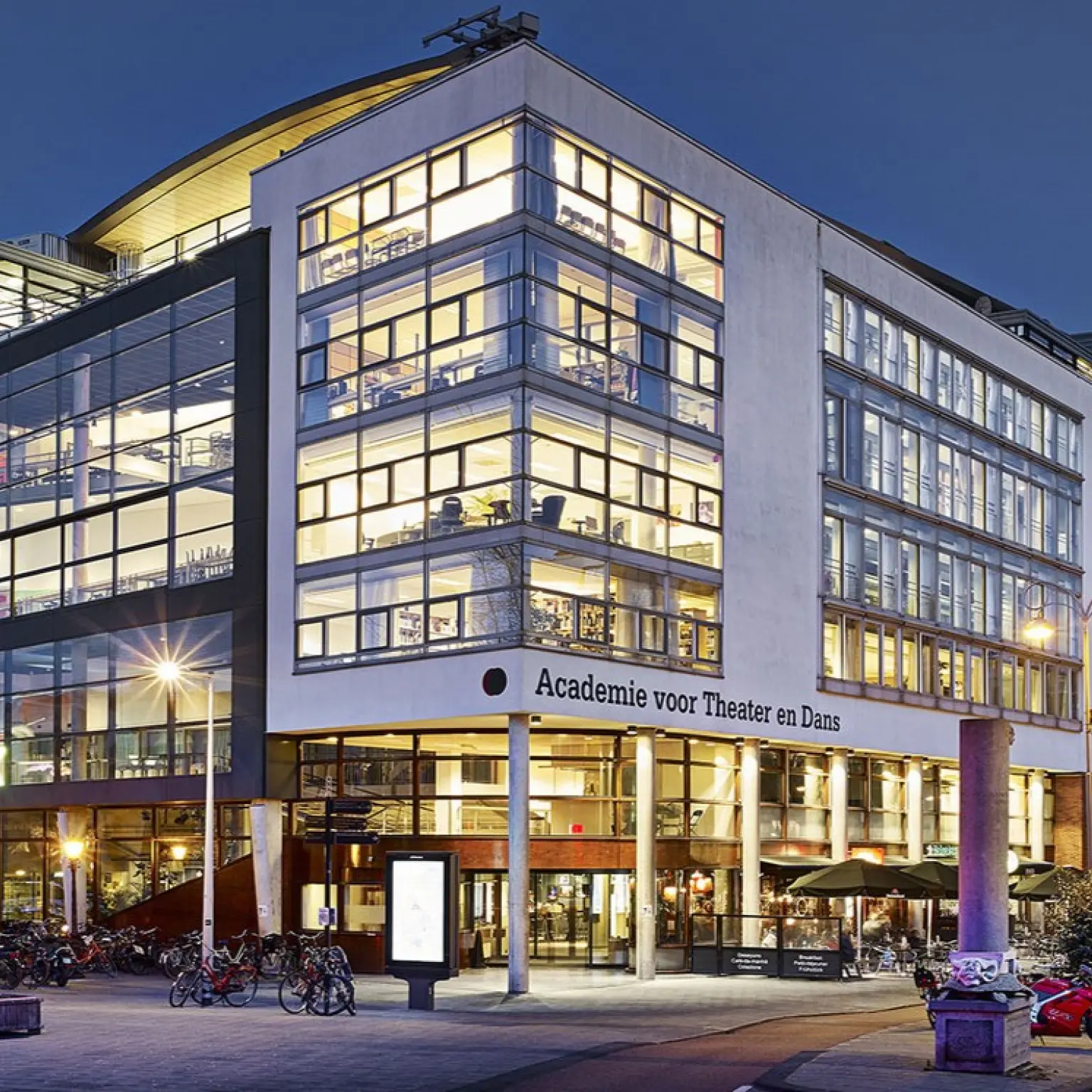 Modern gebouw van de Academie voor Theater en Dans, gefotografeerd in de schemering. Het meerlaagse pand heeft grote glazen gevels waardoor verlichte les- en werkruimtes zichtbaar zijn. Voor de entree staan fietsen geparkeerd en branden straatlantaarns. Op de gevel staat de tekst “Academie voor Theater en Dans”.