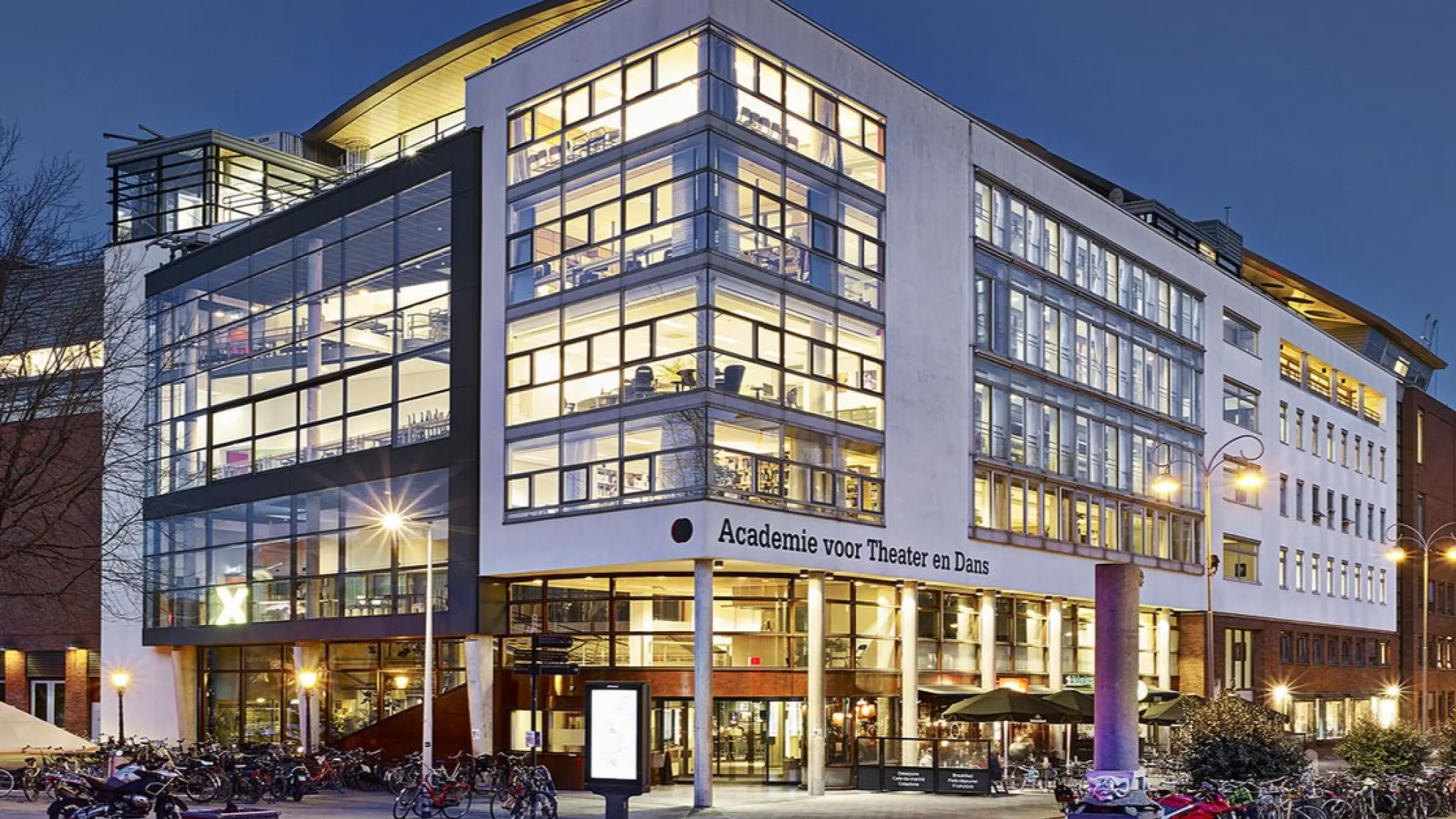 Modern gebouw van de Academie voor Theater en Dans, gefotografeerd in de schemering. Het meerlaagse pand heeft grote glazen gevels waardoor verlichte les- en werkruimtes zichtbaar zijn. Voor de entree staan fietsen geparkeerd en branden straatlantaarns. Op de gevel staat de tekst “Academie voor Theater en Dans”.