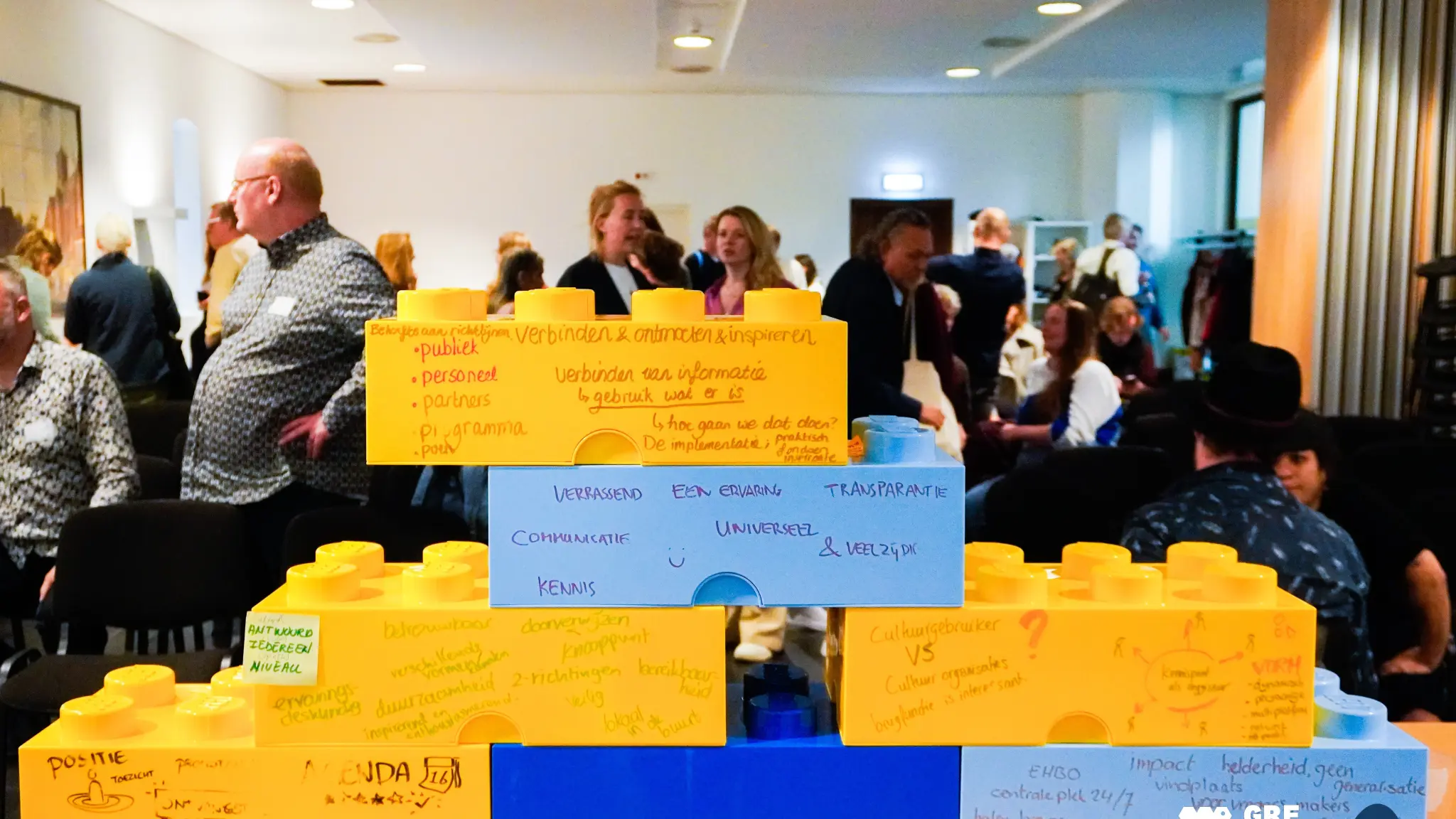 Grote gekleurde bouwstenen met handgeschreven ideeën staan centraal tijdens een interactieve bijeenkomst, terwijl deelnemers op de achtergrond met elkaar in gesprek zijn.