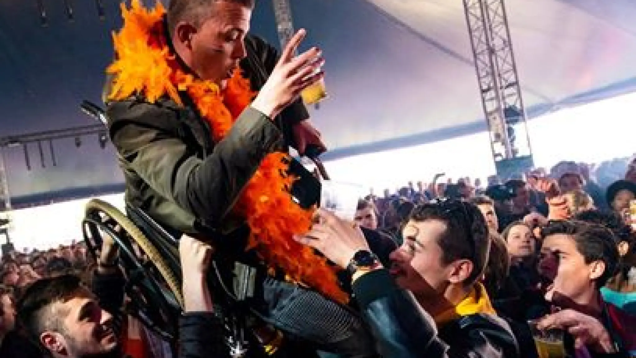 Een jongen in een rolstoel is aan het crowdsurfen bij een Koningsdagfestival. Hij draagt een glas bier in zijn hand, heeft een Nederlandse vlag op zijn wang getekend en draagt een oranje boa.