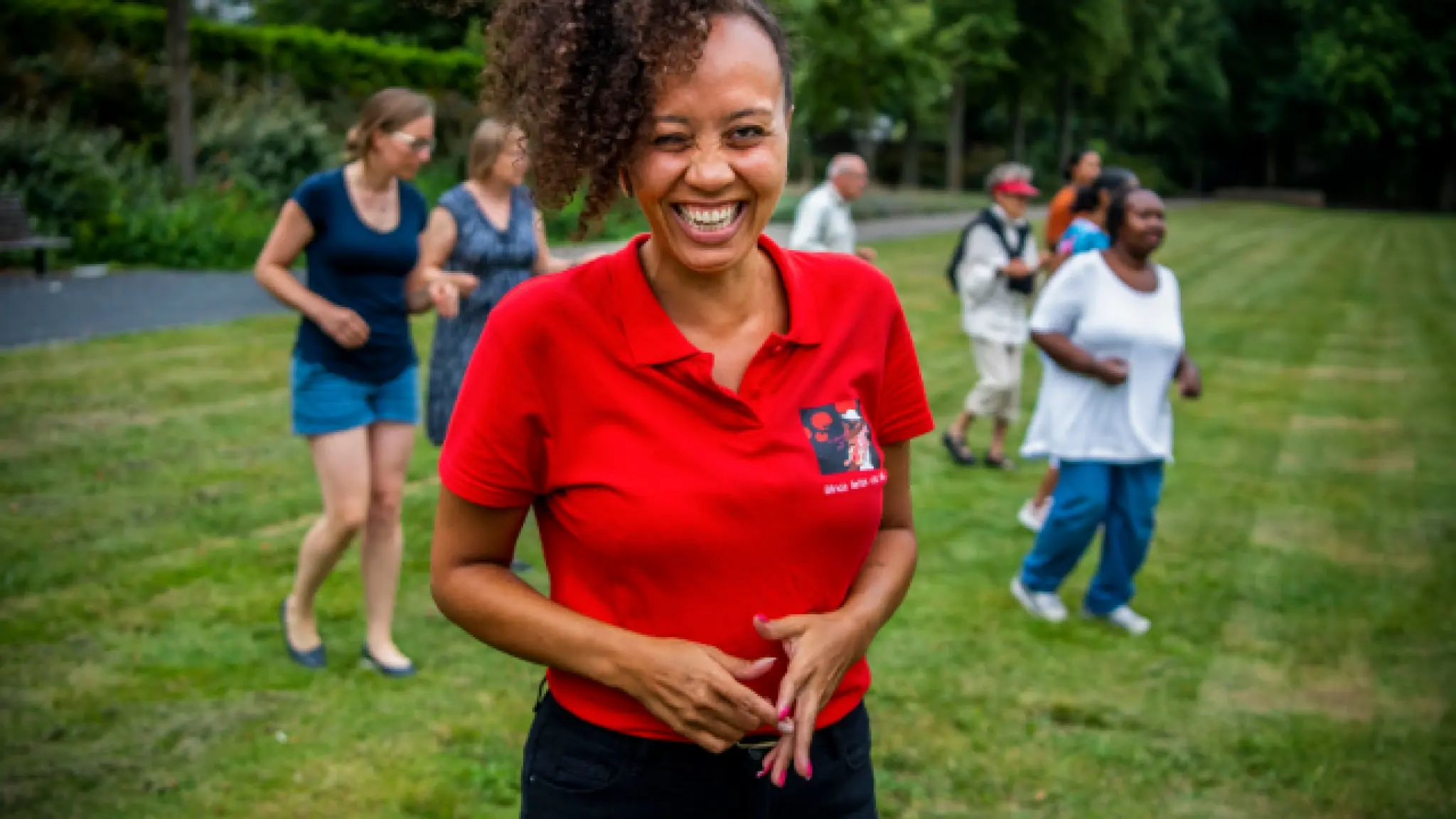Een lachende persoon in een rood poloshirt staat op een grasveld in een parkachtige omgeving. Op de achtergrond zijn meerdere mensen zichtbaar die verspreid over het veld staan of lopen.