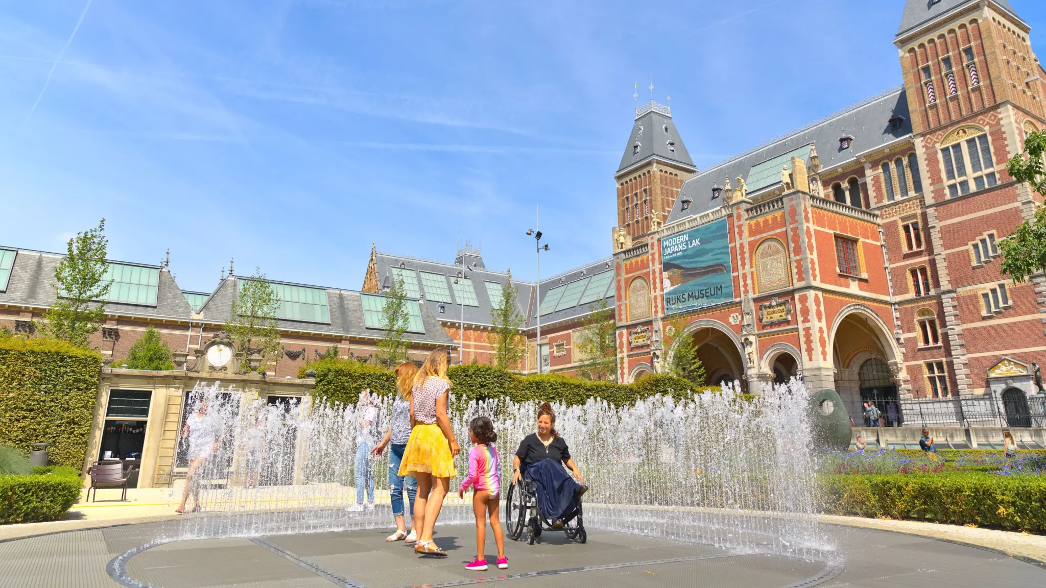 Kinderen spelen in de fontein voor het Rijksmuseum. Ook een vrouw in een rolstoel staat middenin de fontein.