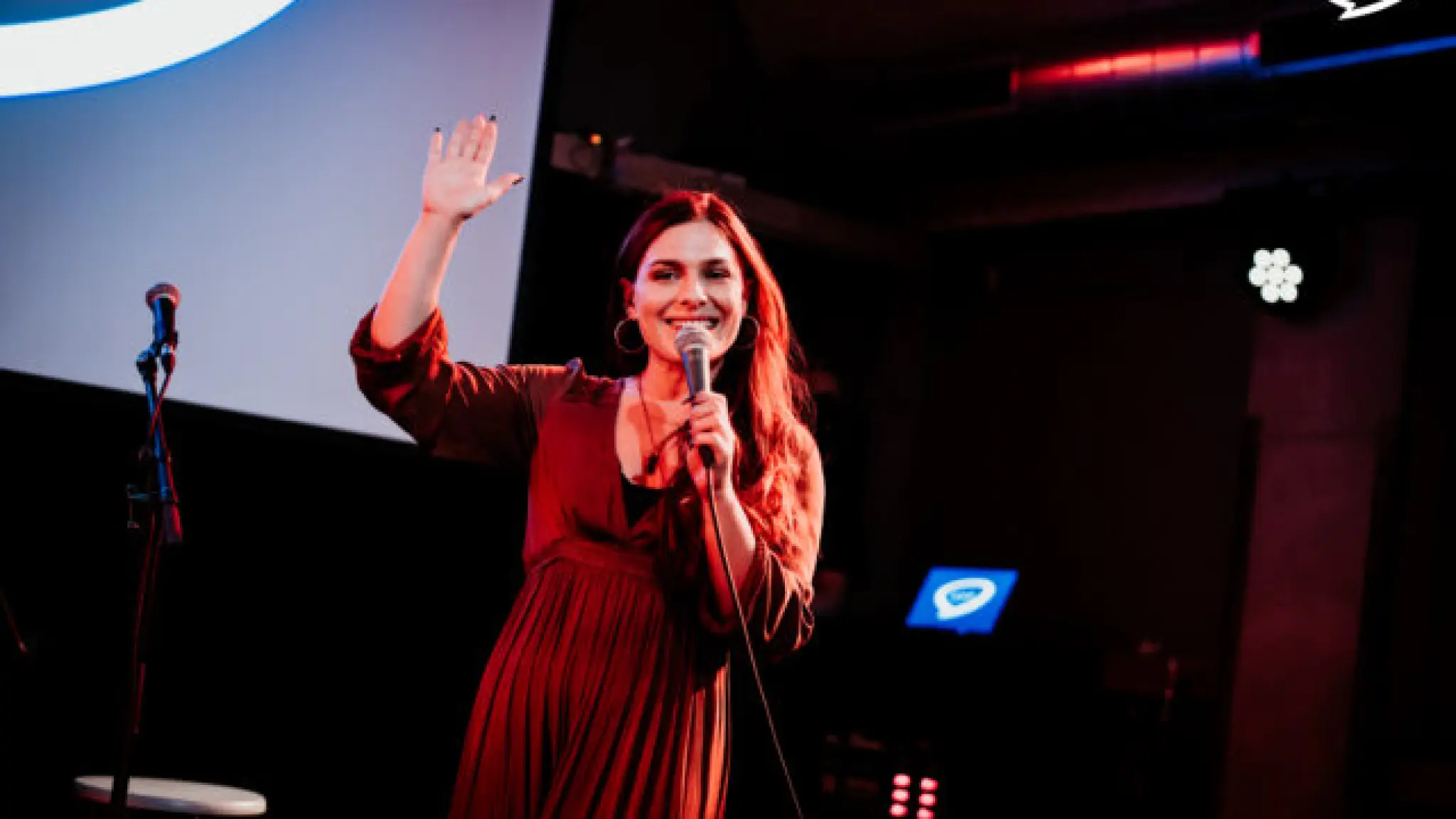 Een persoon in een rode jurk staat op een podium, spreekt in een microfoon en zwaait glimlachend naar het publiek. Op de achtergrond zijn een projectiescherm en podiumverlichting te zien.