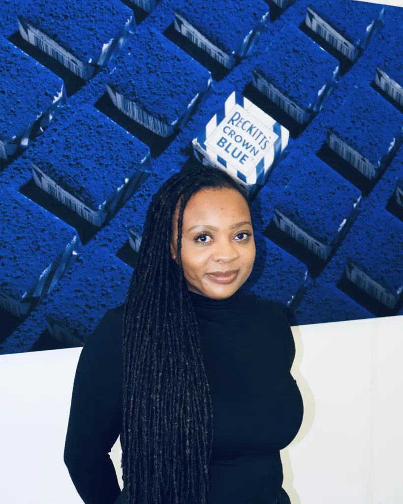 Cheryl Mak heeft haar haar in lange dreadlocks of vlechtjes en ze draagt een zwarte coltrui. Ze staat voor een grote afbeelding van blauwe blokken met op een ervan de tekst Reckitt's Crown Blue. Ze kijkt recht in de camera en glimlacht licht."