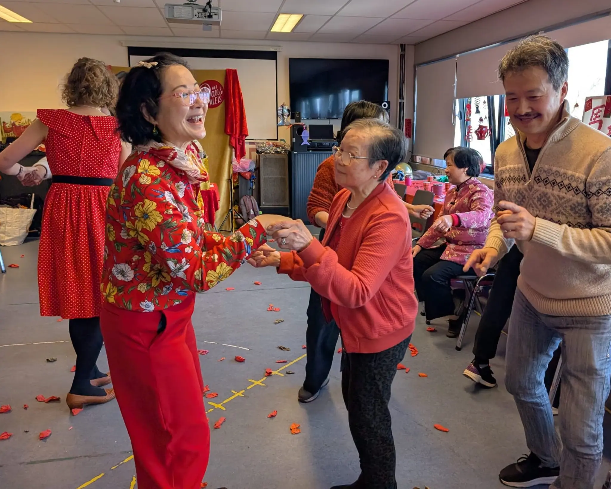 Een groep mensen danst samen in een lichte ruimte. Op de voorgrond dansen twee mensen tegenover elkaar en houden elkaars handen vast, terwijl een derde persoon naast hen meebeweegt.