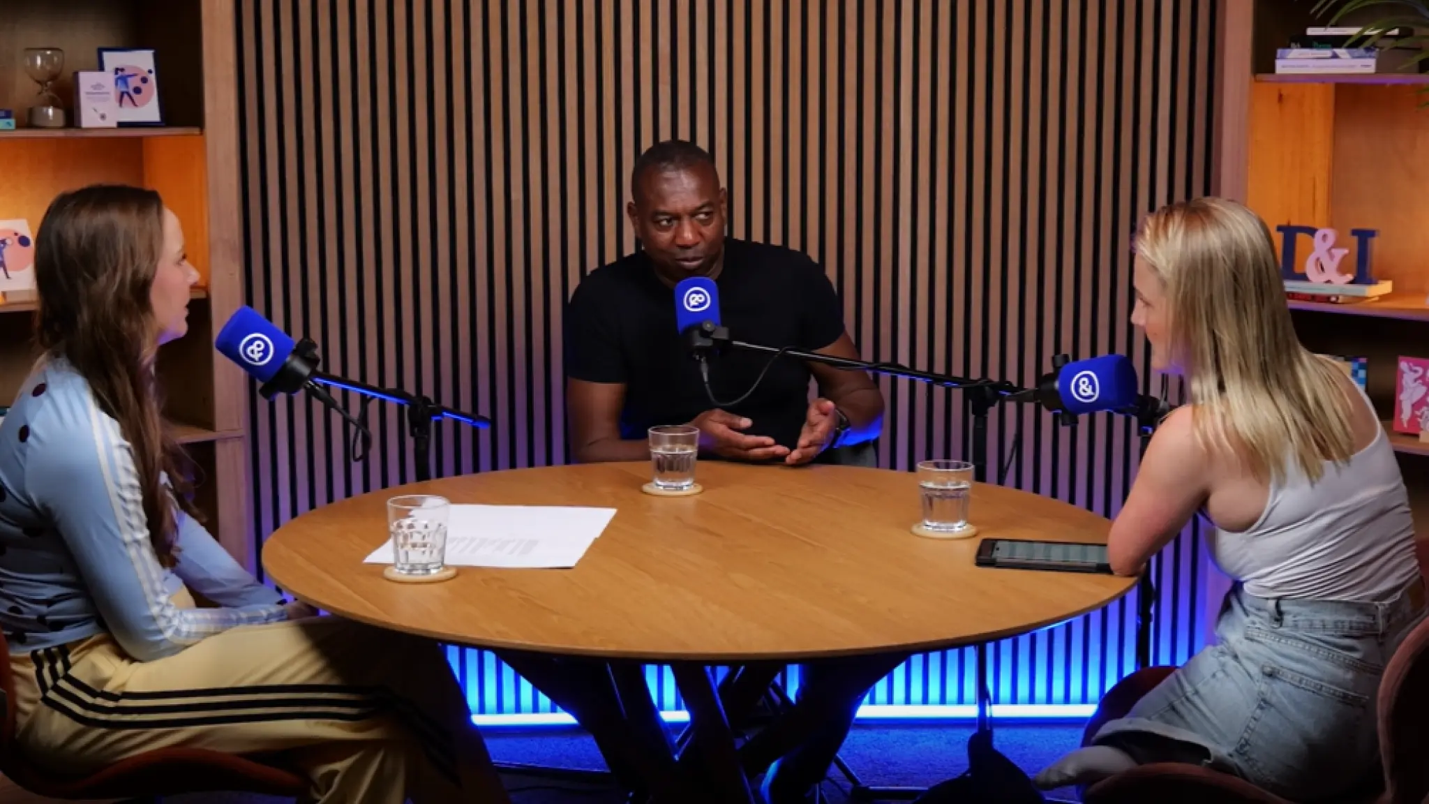 Drie personen zitten aan een ronde tafel in een podcaststudio en voeren een gesprek achter microfoons. Op tafel staan glazen water, een vel papier en een tablet. De studio heeft een houten achtergrond met sfeerverlichting.