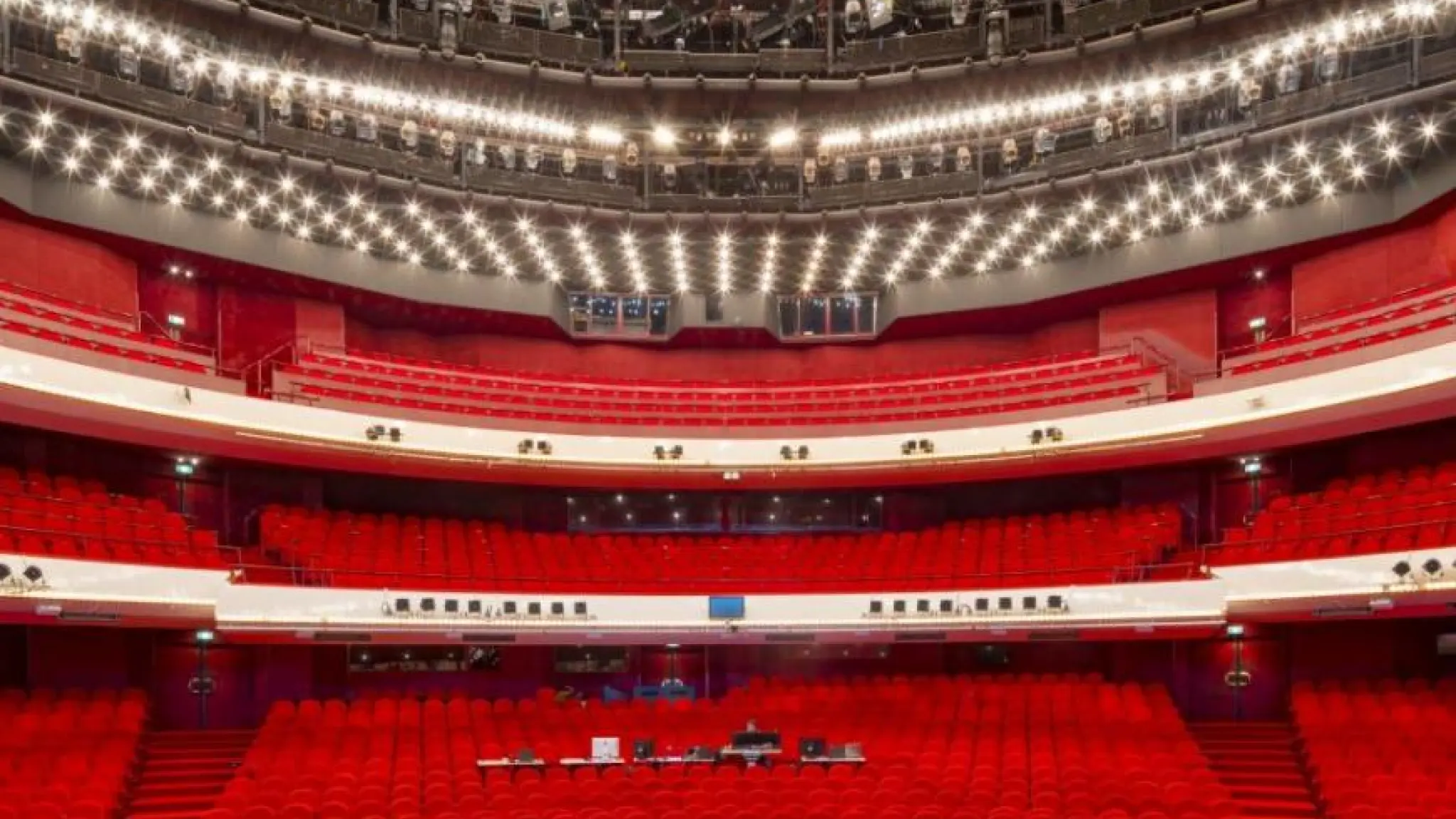 Een groot, modern theater met felle rode stoelen, meerdere balkonlagen en een plafond vol heldere lampen en technische apparatuur. Het zicht is gericht op het publiek vanuit het perspectief van het podium.