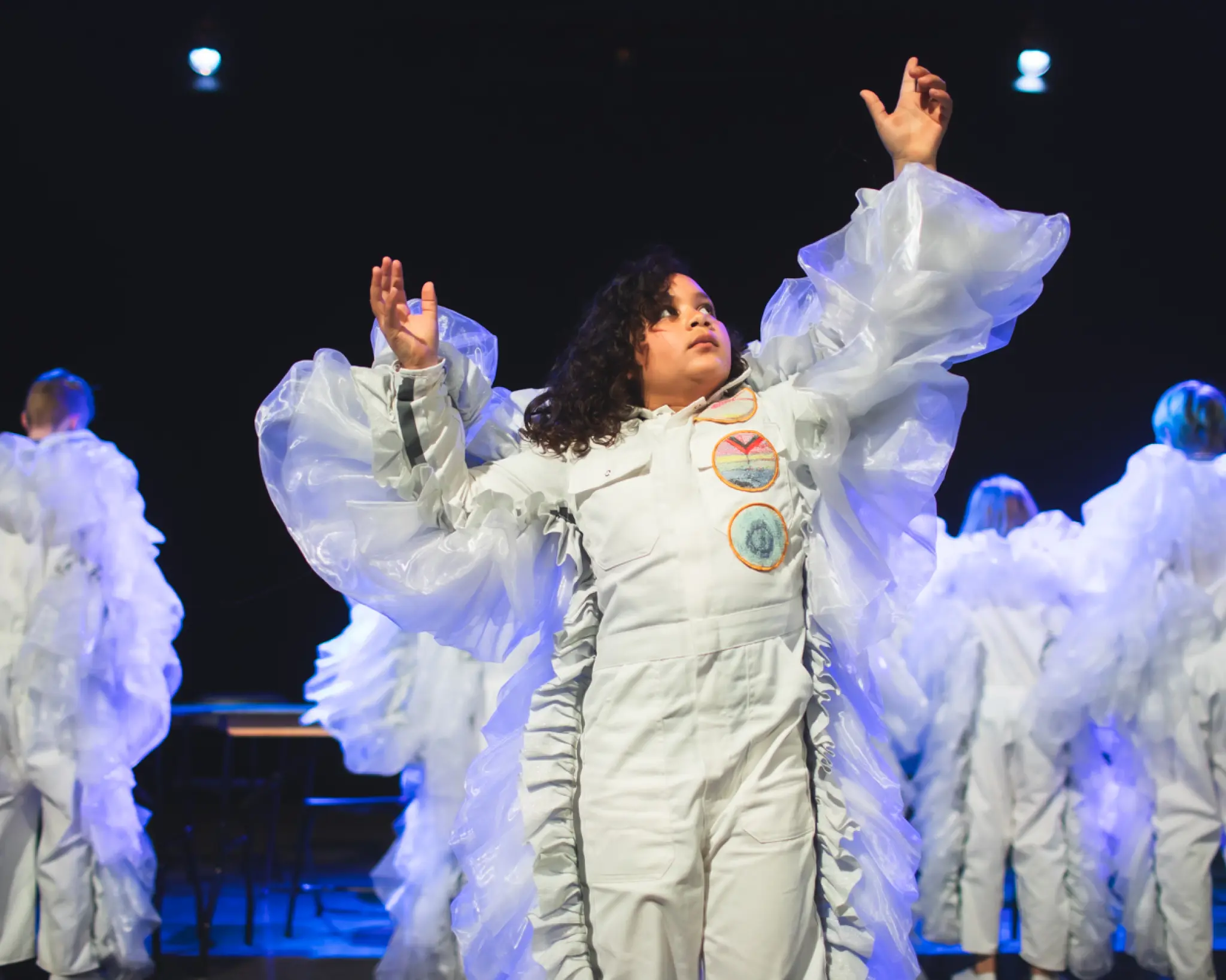 Op een donker theaterpodium staat een performer centraal in een wit kostuum met grote, transparante ruches langs de armen en zijkanten. Met beide armen opgeheven kijkt de persoon omhoog, alsof die een dans- of theatervoorstelling uitvoert. Op de achtergrond staan meerdere performers in vergelijkbare witte kostuums, deels onscherp en met de rug naar het publiek. Blauwe podiumverlichting benadrukt de dromerige, bijna buitenaardse sfeer van de scène.
