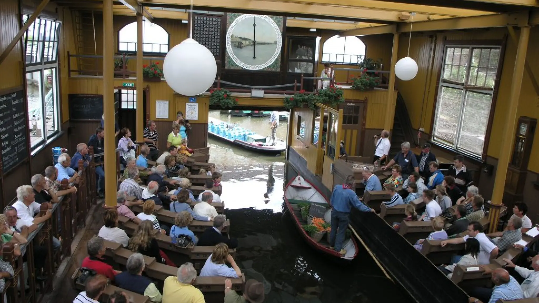 Demonstratieveiling in Museum Broekerveiling. EEn schipper met wortels in zijn boot vraart door het kanaaltje in het midden van het veilinggebouw. Links en rechts van het kanaaltje zitten bezoekers in een soort kerkbanken. Boven de ingang van het kanaaltje hangt een veilingklok. Vooraan staat de veilingmeester met een microfoon in zijn hand.