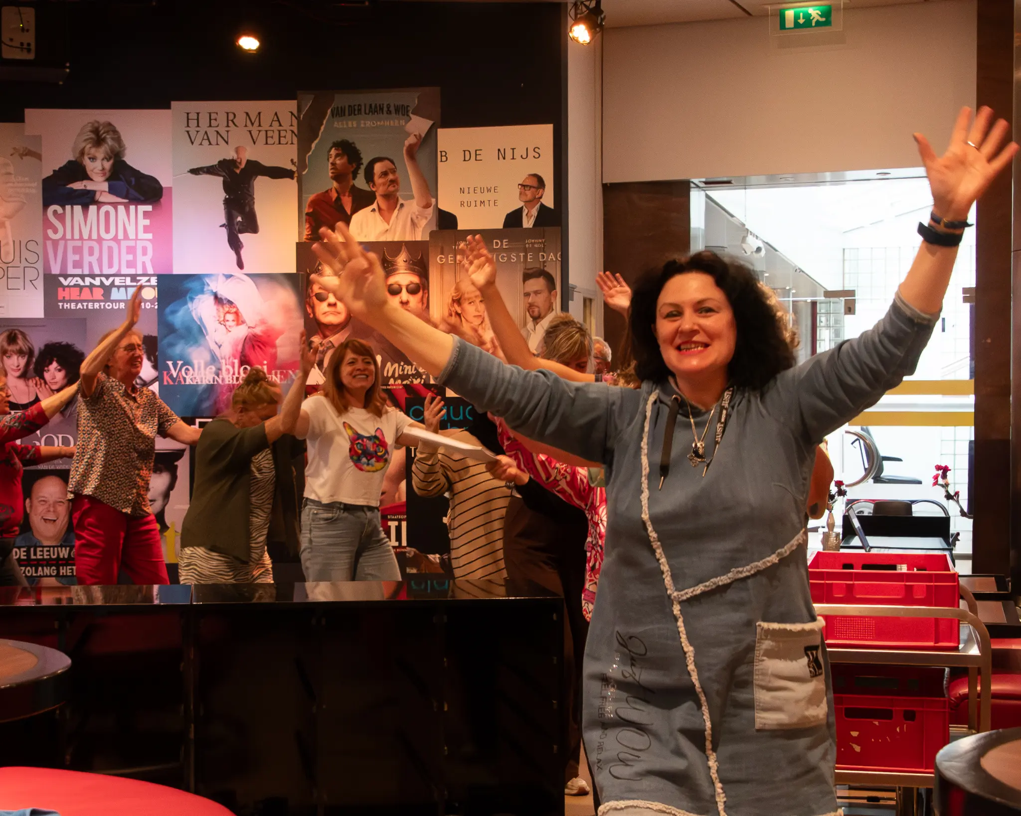 Een groep mensen loopt de polonaise door de foyer van een theater. Op de muur achter hen zijn posters van theatervoorstellingen te zien.