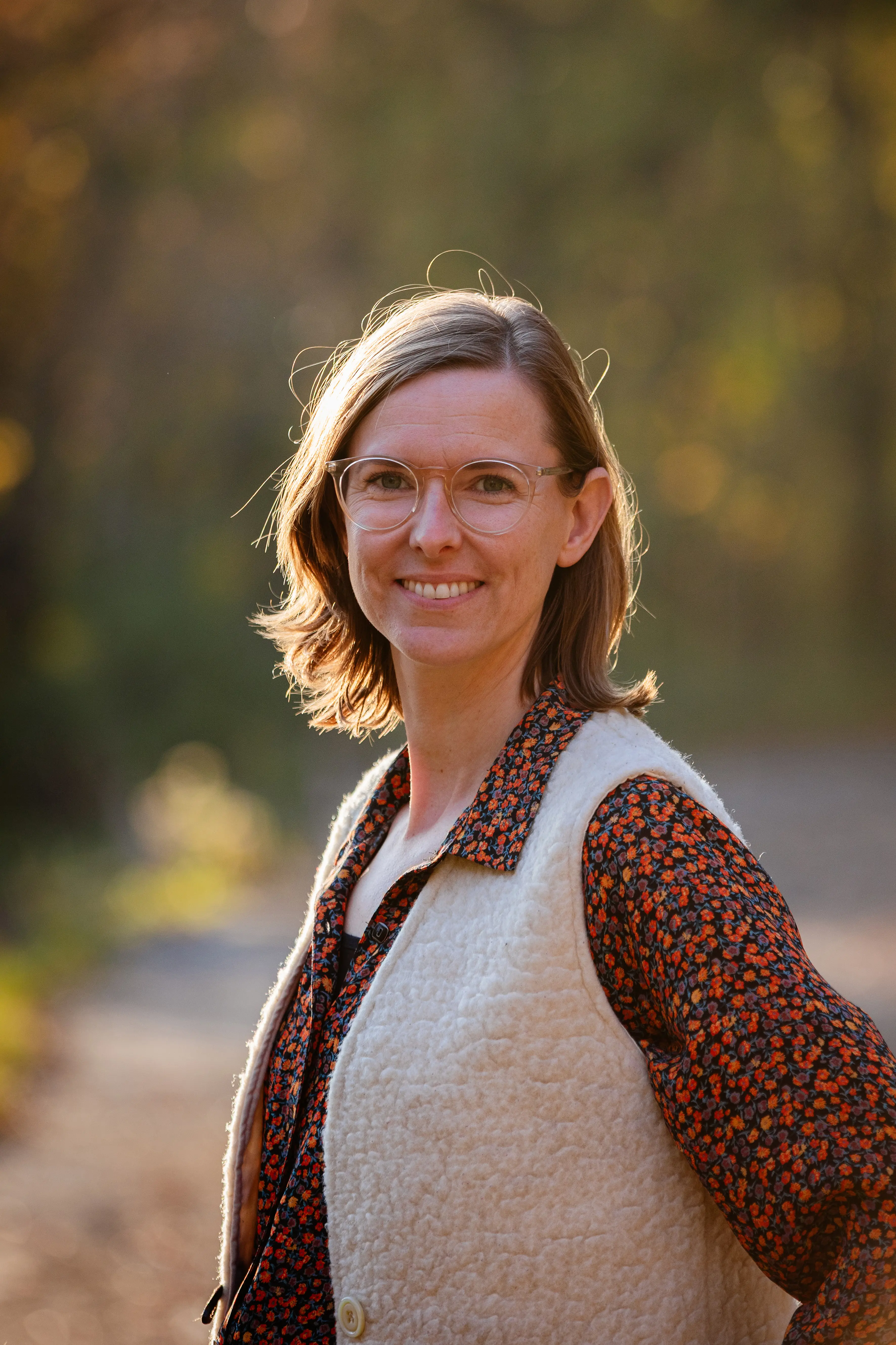 Roos Wattel Draagt een blouse met rode bloemetjes en een wit vest. Ze is gefotgrafeerd op een pad in een park of bos. Op de achtergrond zijn onscherp bomen te zien. Het zonlicht schijnt door de bomen op haar hoofd. Ze glimlacht naar de camera.