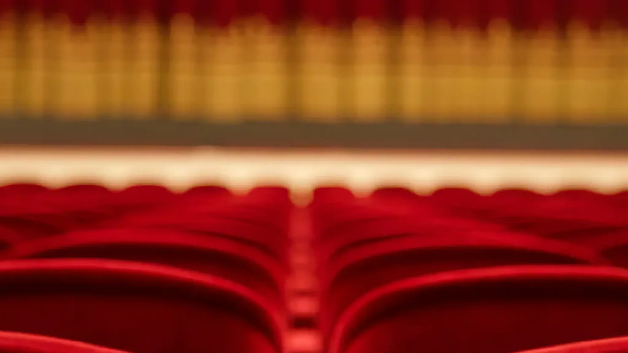 De achterkanten van tientallen rode theaterstoelen, met in de verte een podium.