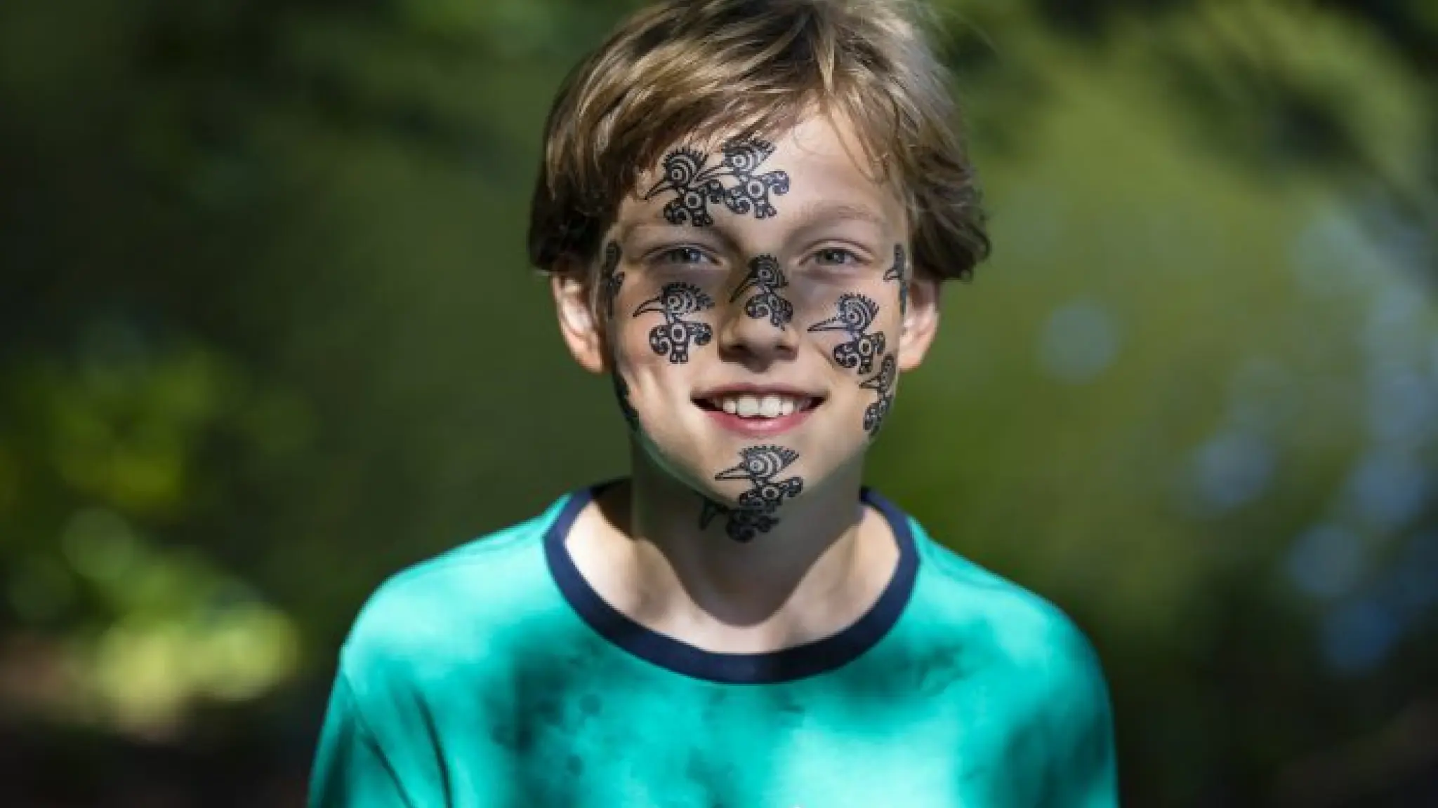 Een jongen in een blauw t-shirt kijkt l;ached naar de camera. Hij heeft diverse tekeningen van vogels op zijn gezicht.