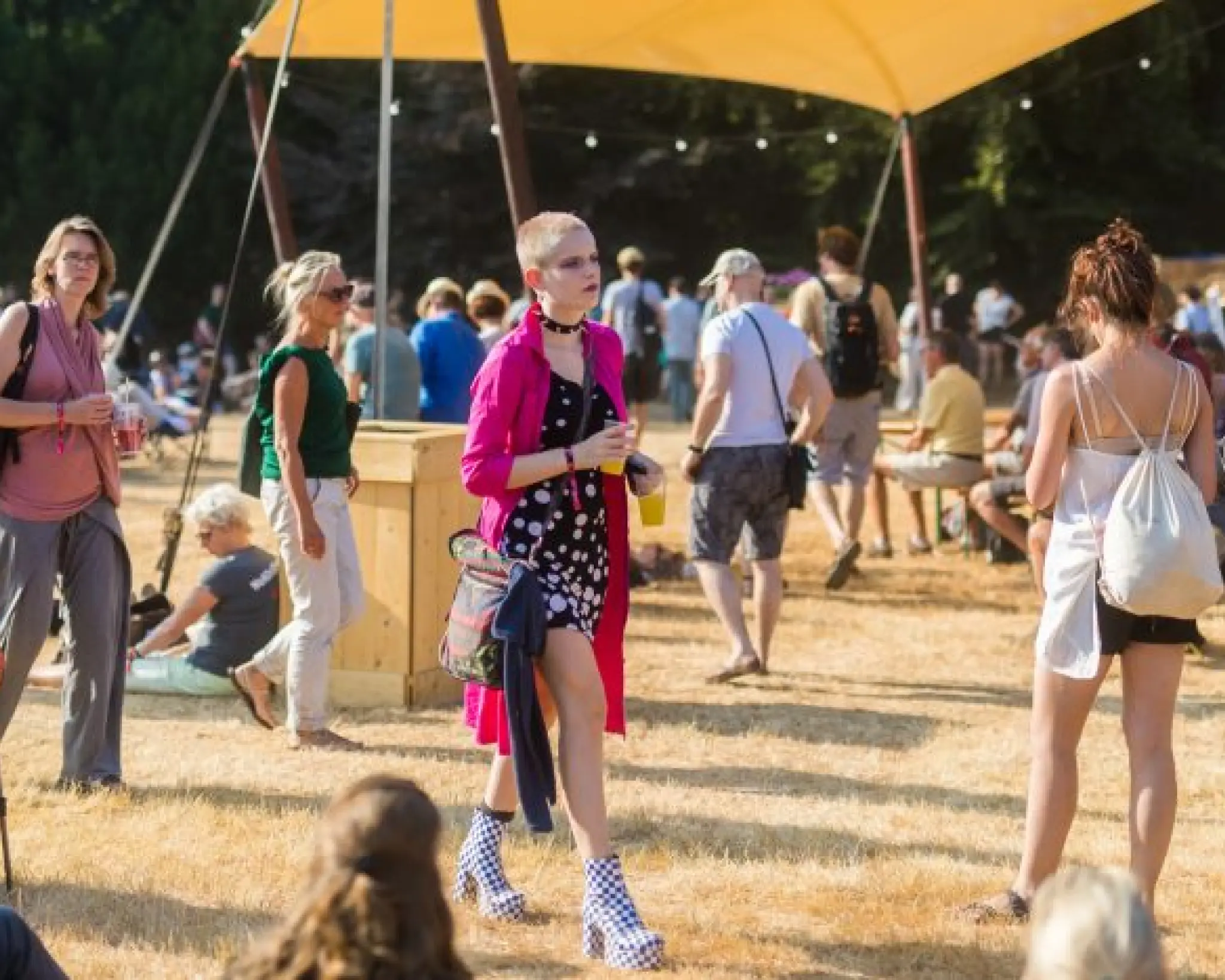 Festivalbezoekers lopen over een door de zon geel geworden grasveld. In het midden staat een geel doek dat tegen de zon gespannen is. In het midden loopt een jone vrouw met een lange roze jas en zwart-wit geblokte laarzen.