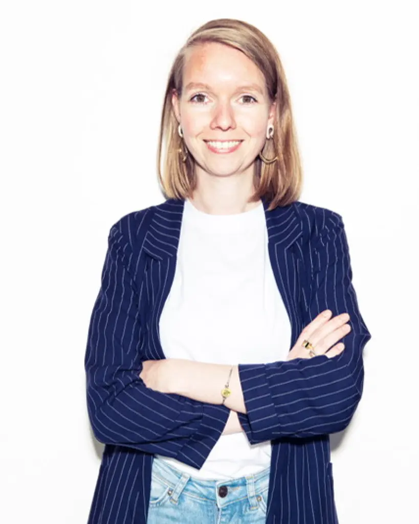Emma staat met haar armen over elkaar recht voor de camera. Ze lacht vriendelijk. Ze draagt een spijkerbroek, wit t-shirt en een blauwe blazer met krijtstreep.