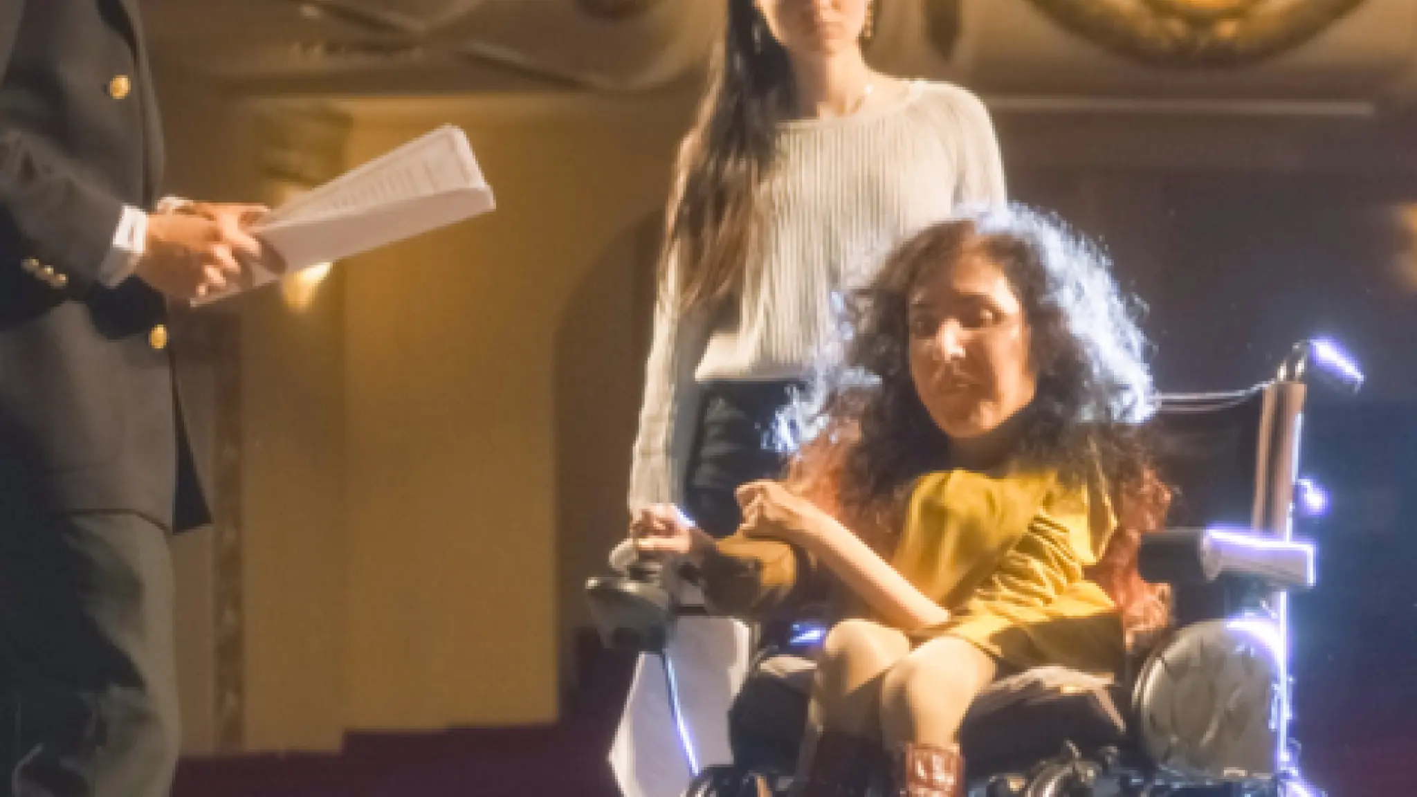 Drie mensen repeteren op een verlicht theaterpodium; een vrouw in een rolstoel staat centraal, begeleid door een andere vrouw, terwijl een man met een script hen aanwijzingen geeft.
